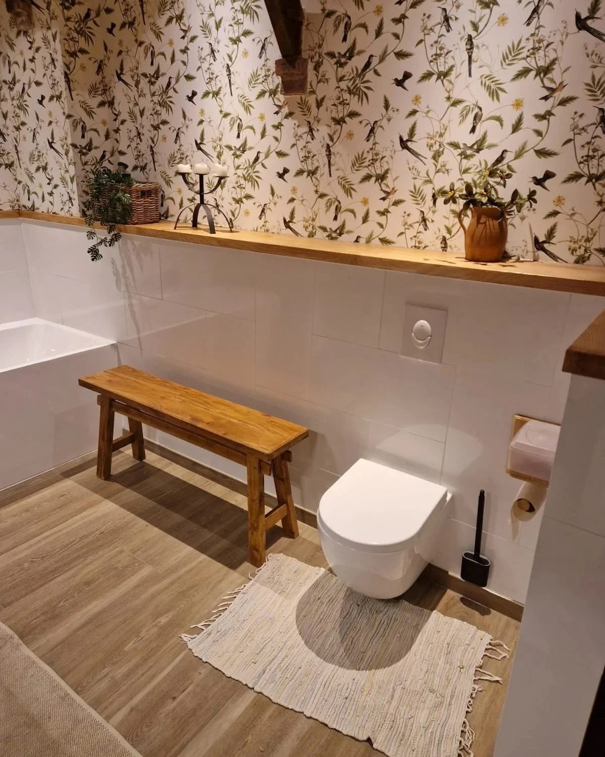 Een badkamer met een geitenbont tapijt, een toilet, een houten bank, een blad met planten en decoratieve items, en een muur met een botanisch behang met vogels en bladeren.