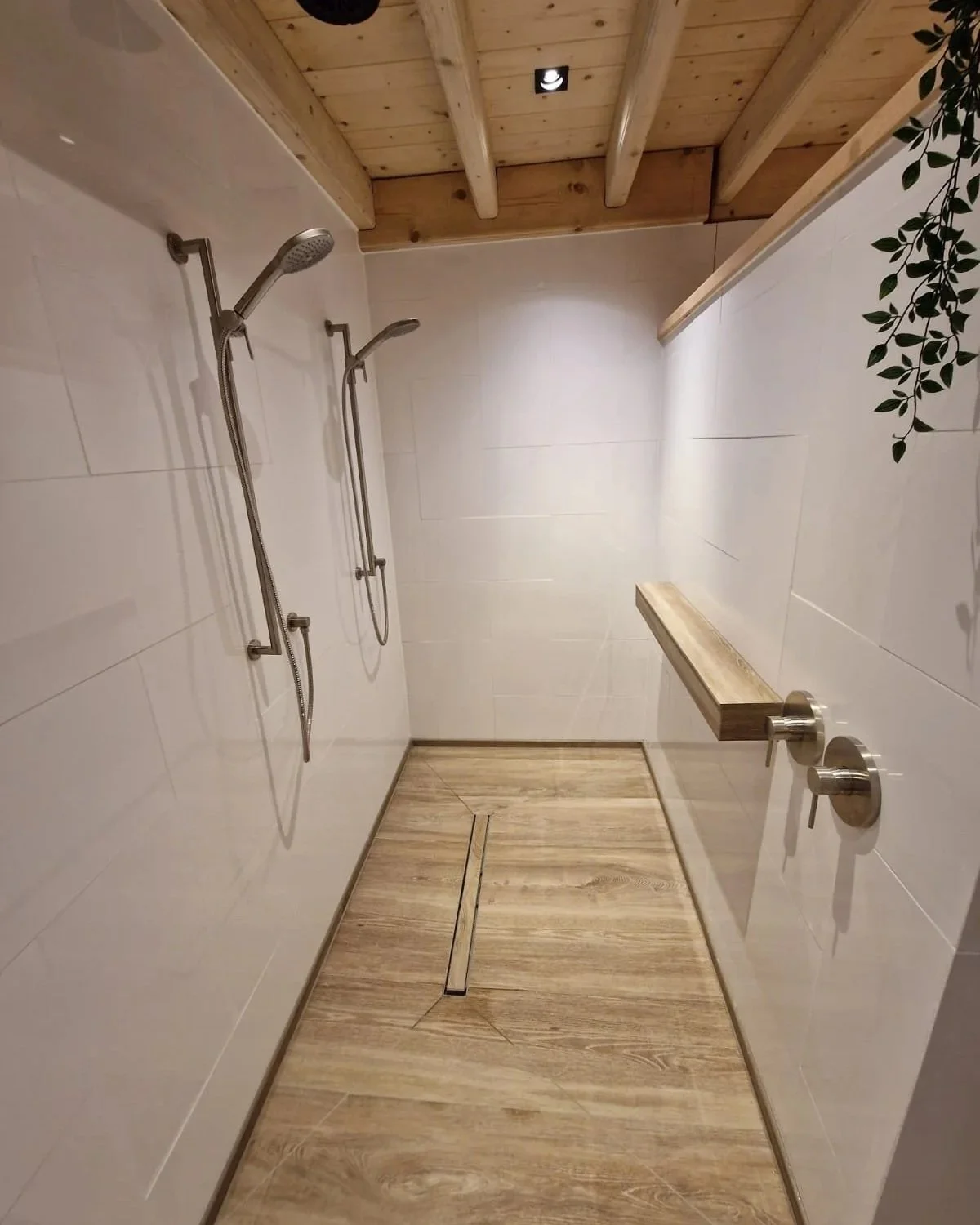 De afbeelding toont een moderne douche met twee douches, een houten vloer en plafond, en eenvoudige muurankers. Er is ook een houten wandje met een plantenrand en twee bedieningsknoppen.