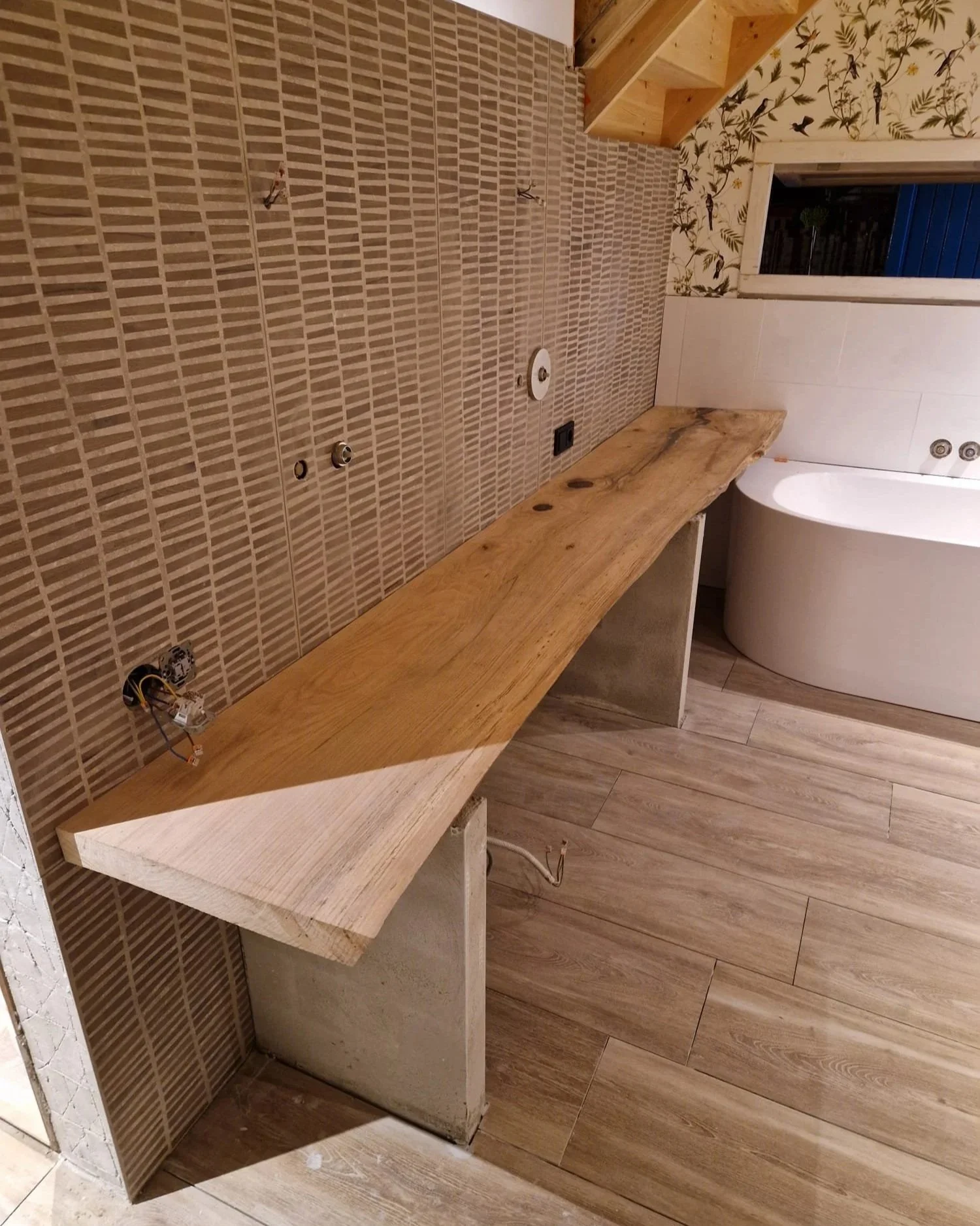 Een badkamer onder renovatie met een houten werkblad of kast die nog afgewerkt moet worden. De muur is gedeeltelijk betegeld met kleine rechthoekige tegels en er zijn openingen voor sanitair en elektriciteit. Er is een badkuip zichtbaar en een raam m