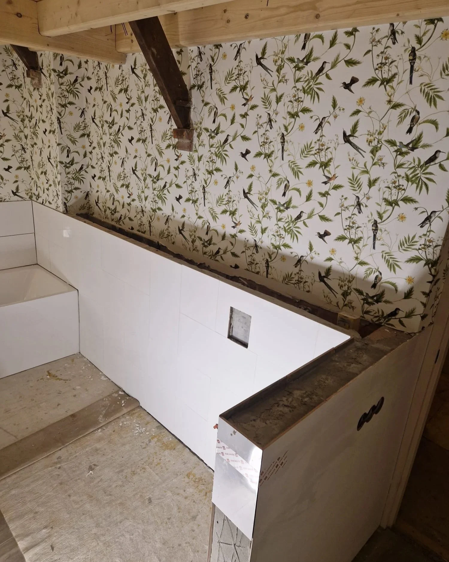 Binnenaanzicht van een badkamer die wordt gerenoveerd, met een betegelde muur met vogel- en bladmotief en een onderbouwkast met een nieuw zwart werkblad.