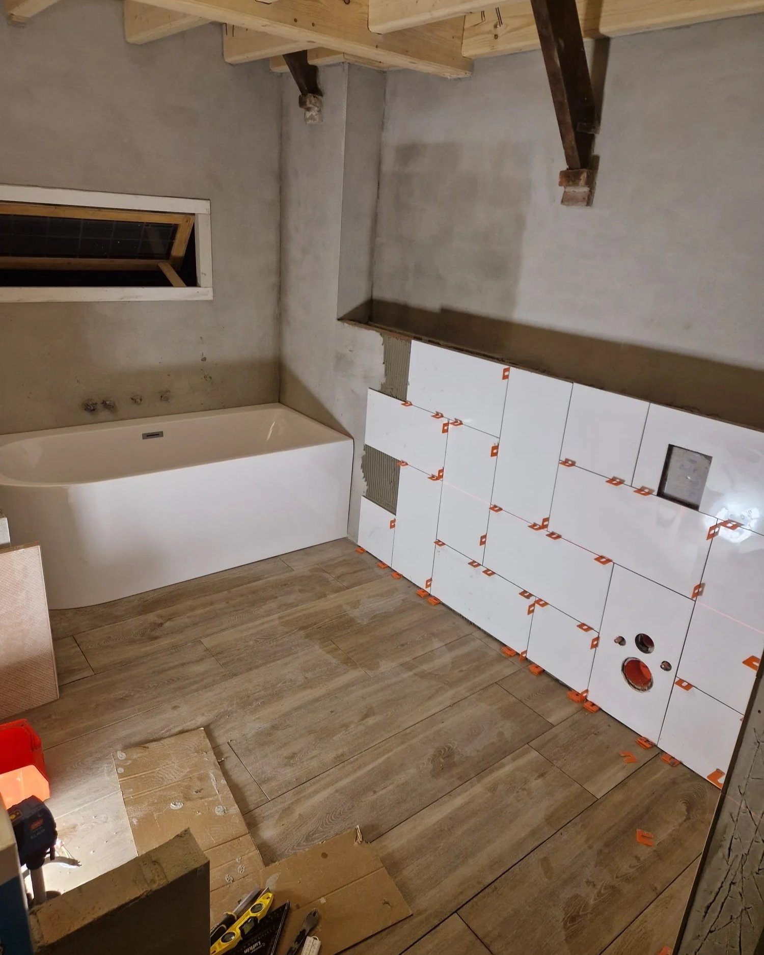 Een badkamer onder renovatie met witte tegels die geïnstalleerd worden, een badkuip, een kleine horizontale raam, en verschillende bouwmaterialen en gereedschap op de vloer.