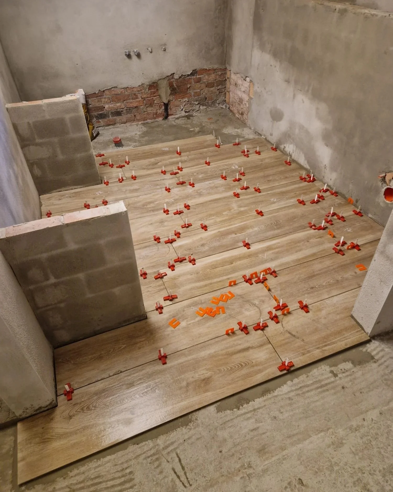 De foto toont een kamer in de bouw of renovatie, met houten vloerplanken die net gelegd worden en vastgezet met rode wiggen. De muren zijn onafgewerkt, met zichtbare bakstenen en beton. Verschillende installatiepunten voor water en elektriciteit zijn