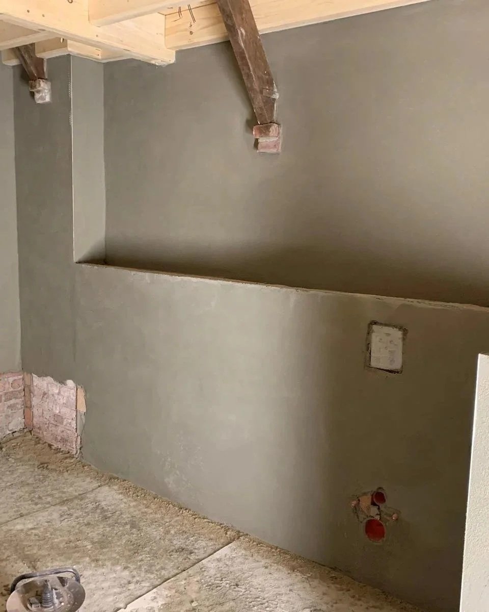 Een geïnstalleerde muur met een geverfde muur en enkele uitsparingen voor elektra en een kabel. Er is een houten plafondbalk en een betonnen vloer.