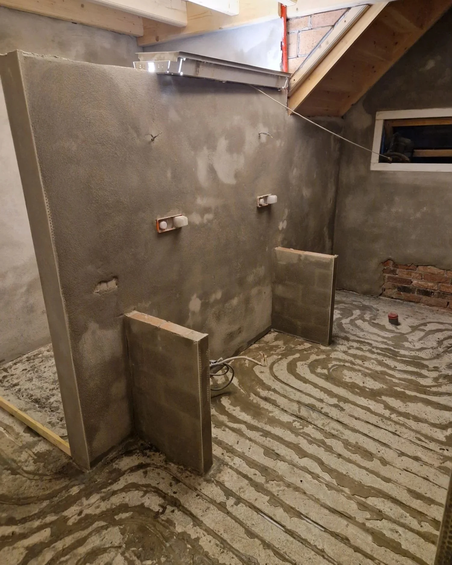 Een kamer onder constructie met een betonnen muur, een tijdelijke wand, verbruikskasten en vloer-rioolbuizen. De vloer en muur zijn onafgewerkt.