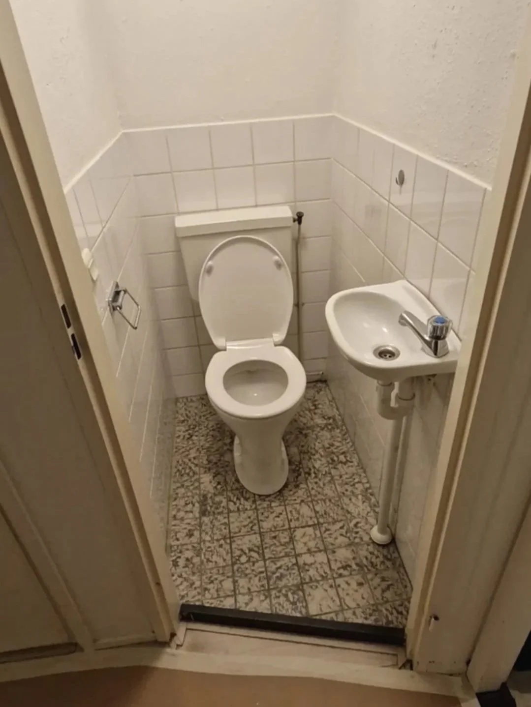 Kleine toiletruimte met een wit toilet en een kleine wit gootstein, witte tegelwanden en een betegelde vloer.
