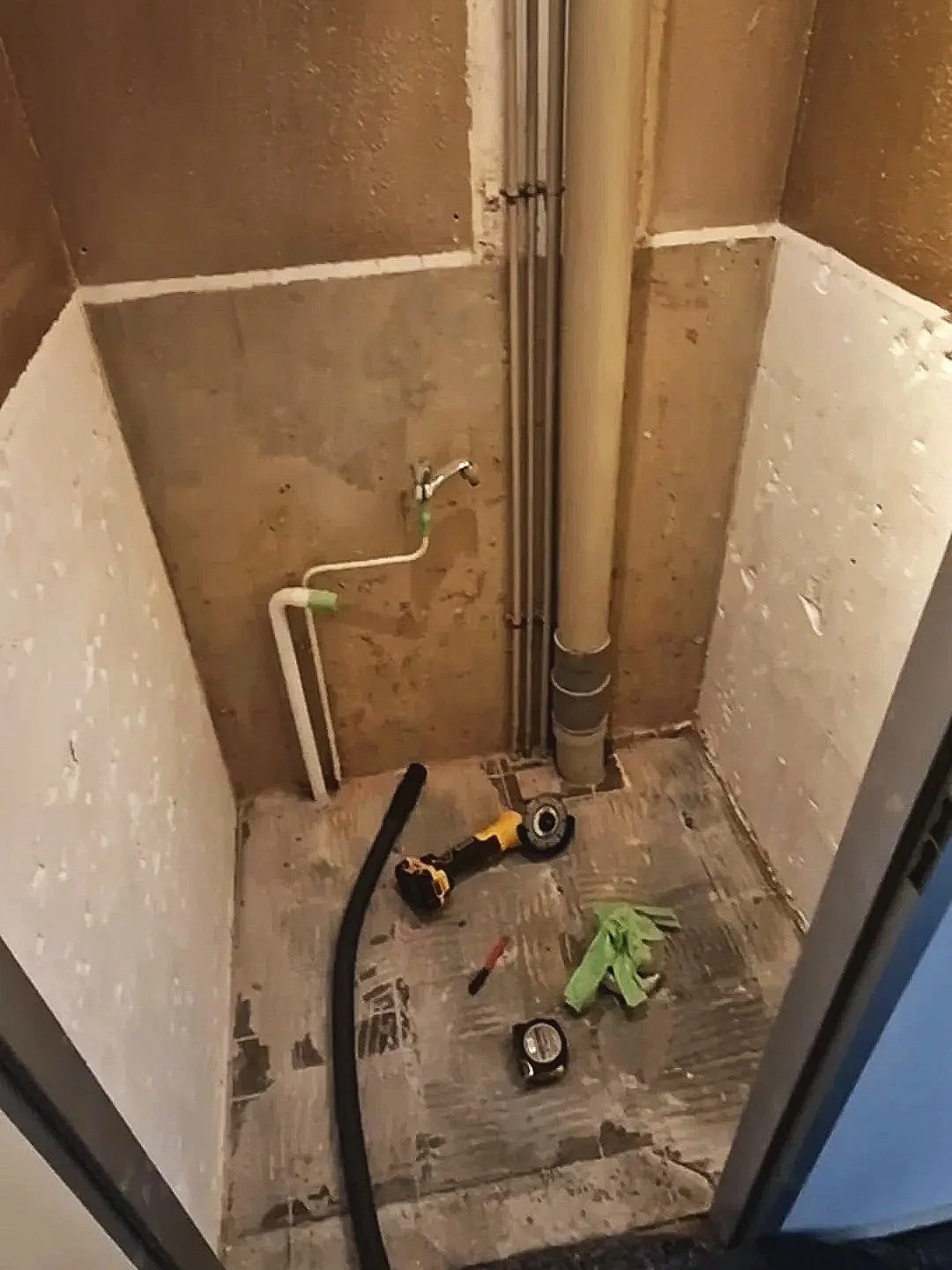 Een kleine werkruimte met bouwmaterialen en gereedschap, inclusief een waterkraan, buizen, een stroomkabel, een tape measure, en een boor op de vloer.