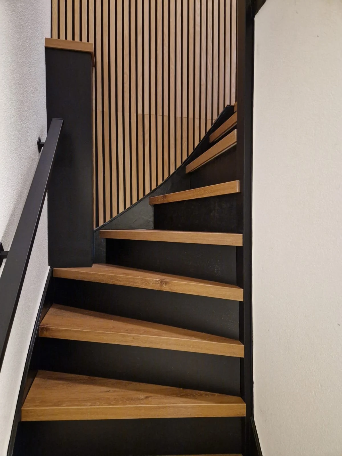 Houten trap met zwarte onderkant en metalen leuning, voorgevel met houten latten en witte muur.