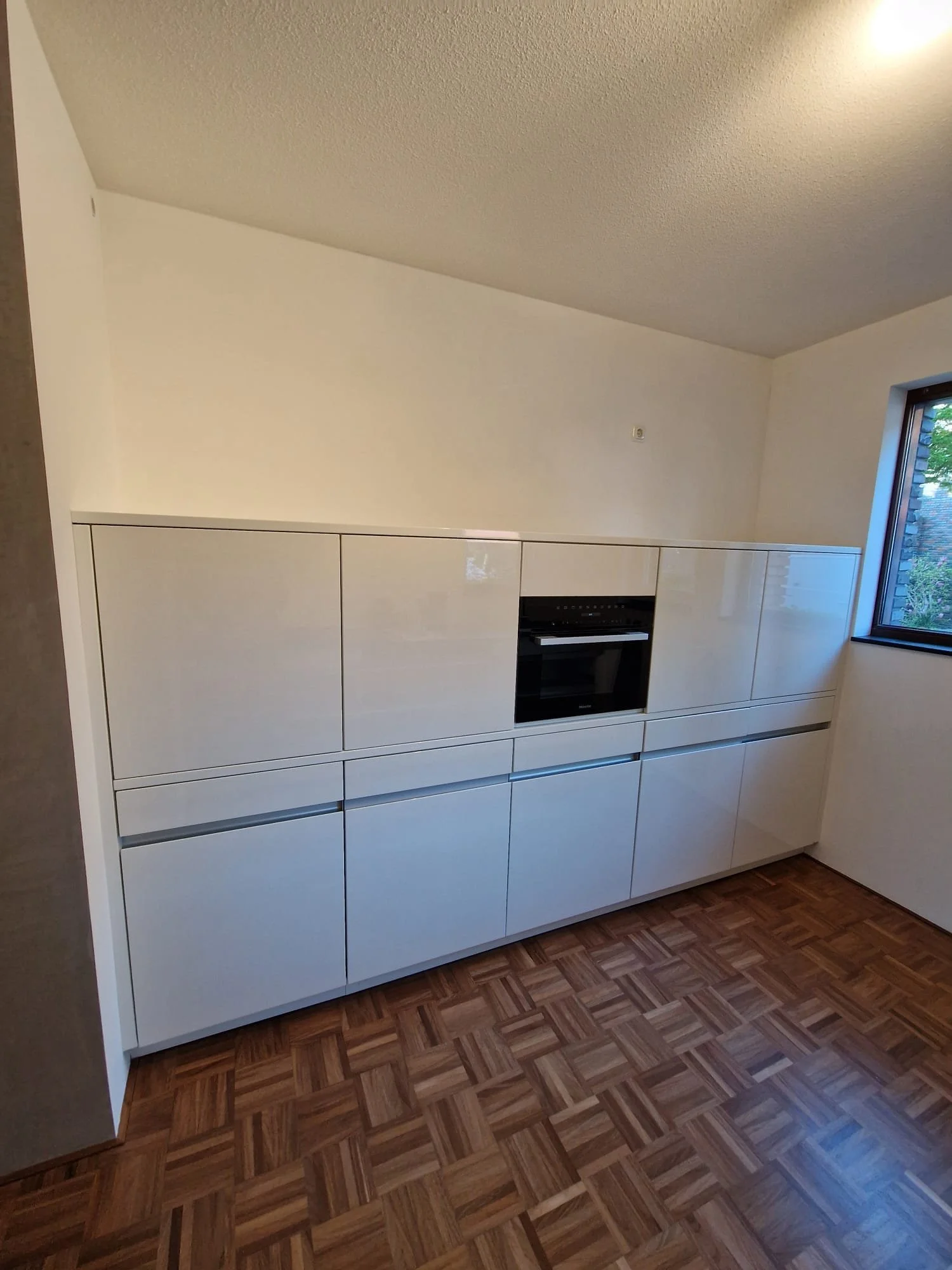 Moderne witte keukenkasten met ingebouwde oven en een raam met uitzicht op buiten.