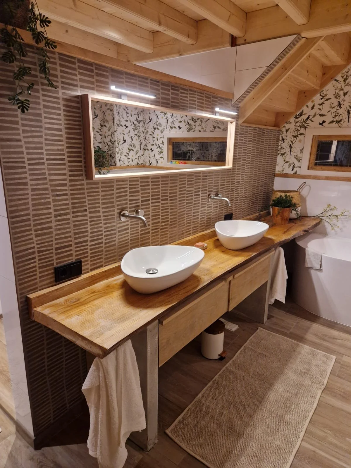 Een badkamer met een houten wastafel met twee witte schaaltjessinkers en een spiegel met verlichting. De muur achter de wastafel is bekleed met bruin geblokt tegels en decoratieve muurpapier met bloemen en vogels. Er zijn ook planten en decoratief mandje op de rand van het blad, er hangt een handdoek onder de wastafel en er is een badkuip met een wit handdoekje.