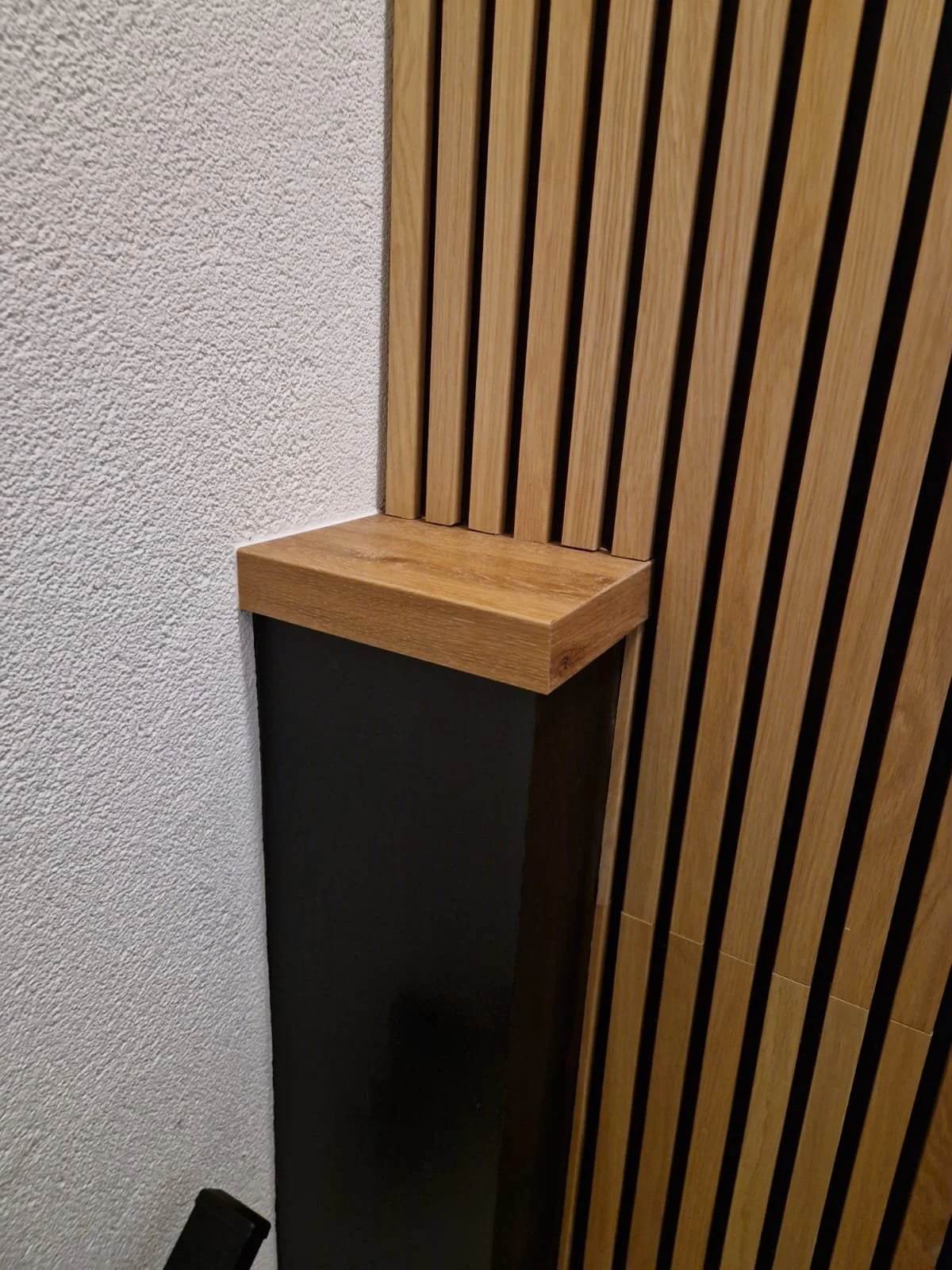 Close-up van een hoek met een houten paneelwand en een zwart onderdeel, met een houten plank op de muur.