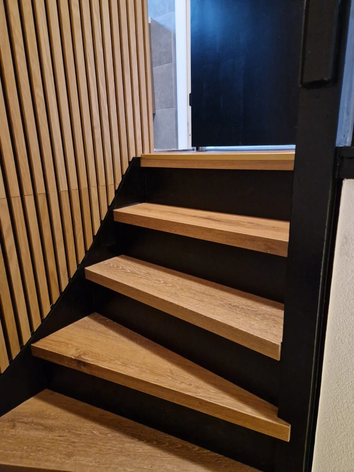 Een trap met houten treden en zwarte zijwanden, naast een muur met houten lamellen en een deur op de achtergrond.