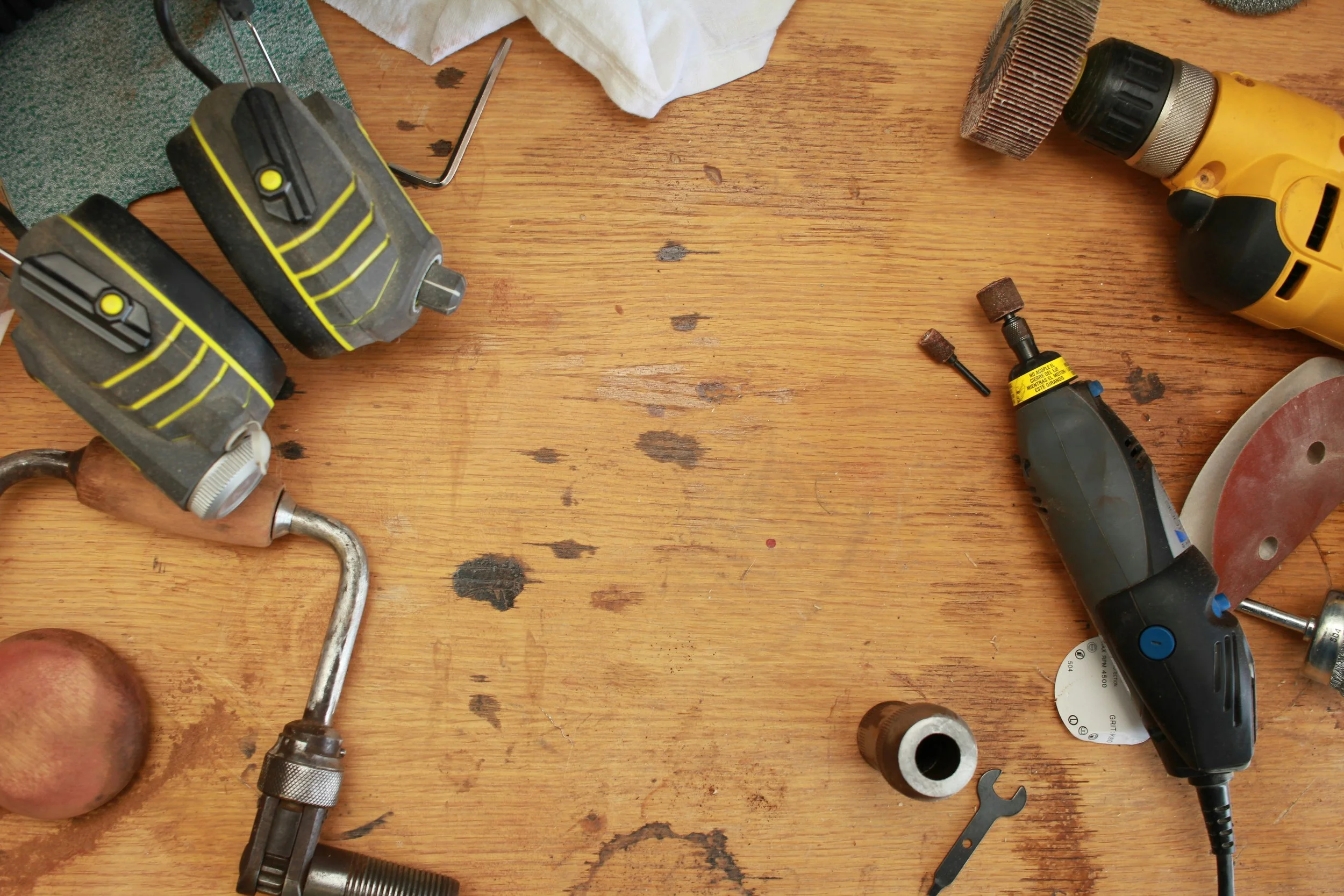 Top-down view of a wooden workbench with various tools and equipment, including drills, a small wrench, a grinding disc, and a power sander, with some oil stains and a potato.