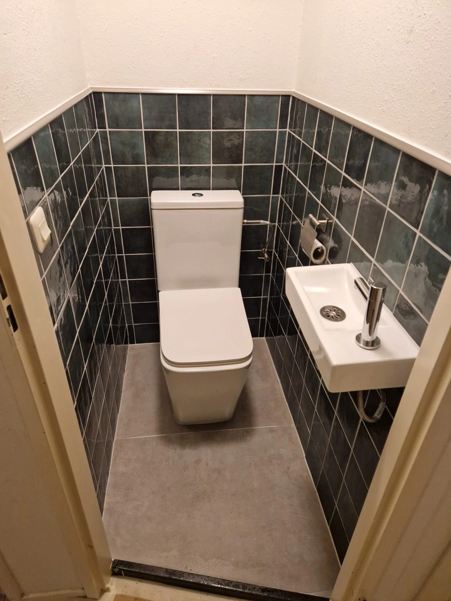 Kleine toilet bevat een toilet en een fontein, met zwart en grijs betegelde wanden en een witte plafond, deurFrame zichtbaar.