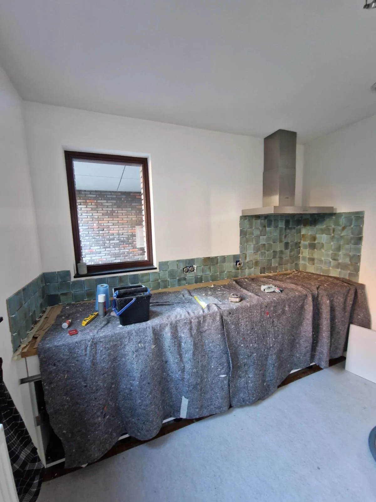 Een keuken onder renovatie met een werkblad bedekt met een grijs werkkleed, groene mozaïek tegels op de muur, en tools en verfbakjes op het werkblad. Er is een raam dat uitkijkt op een bakstenen muur.