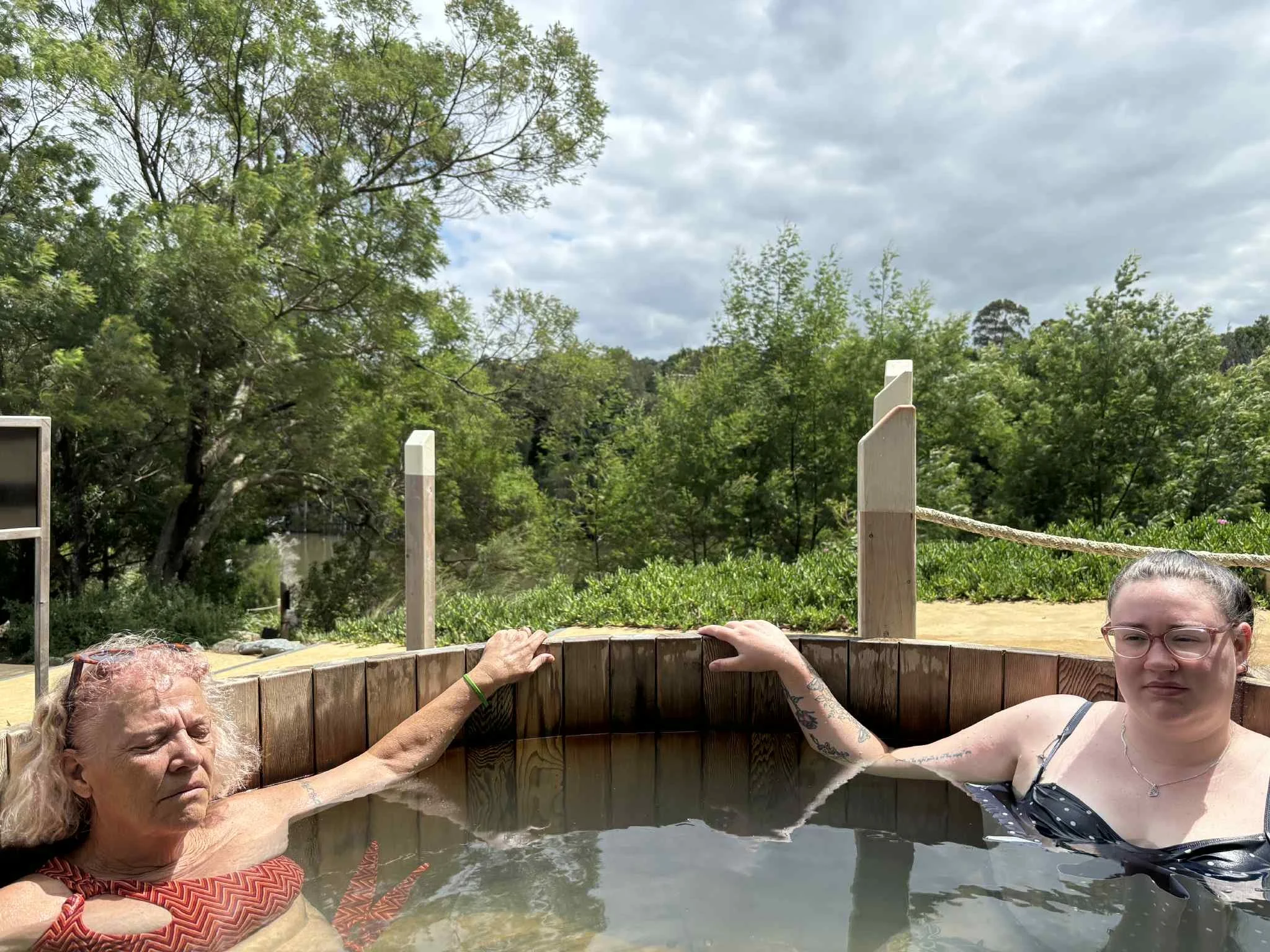 Two women relaxing in a wooden hot tub outdoors, surrounded by green trees and cloudy sky.