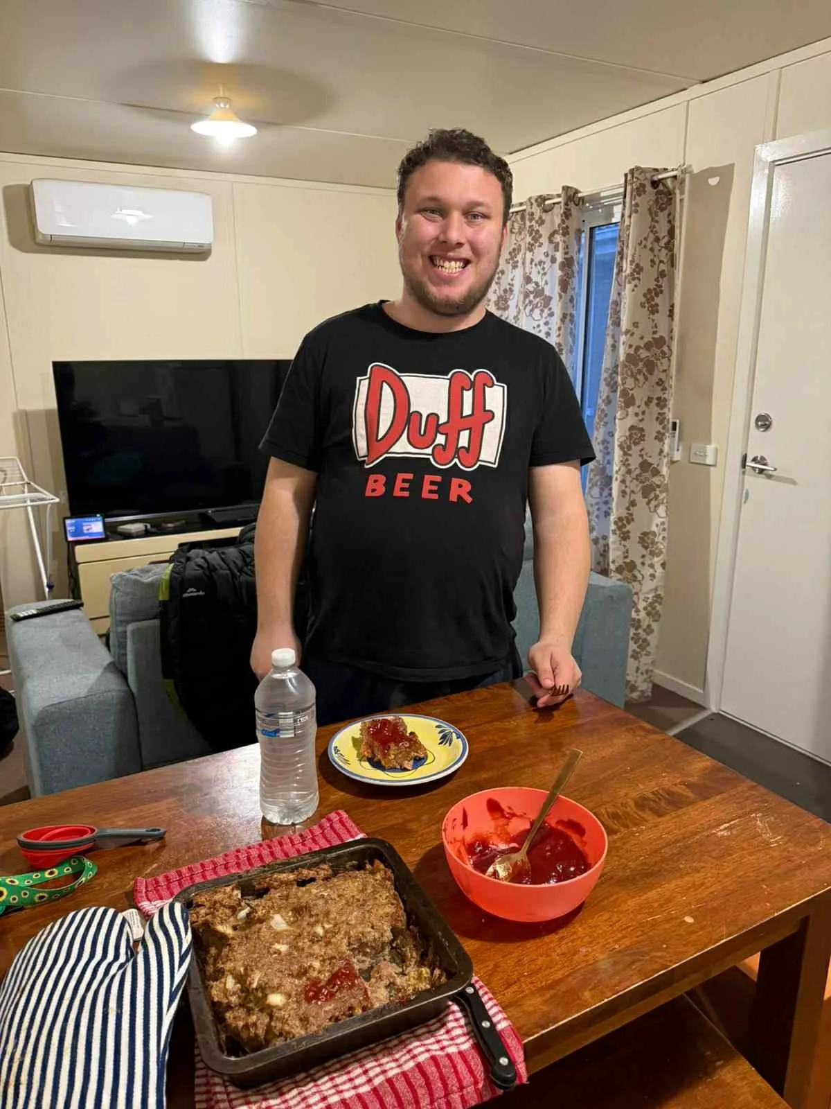 A man smiling at the camera in a living room, standing behind a table with a partially eaten meatloaf, a red bowl of ketchup, a bottle of water, scissors, and a patterned oven mitt on it.