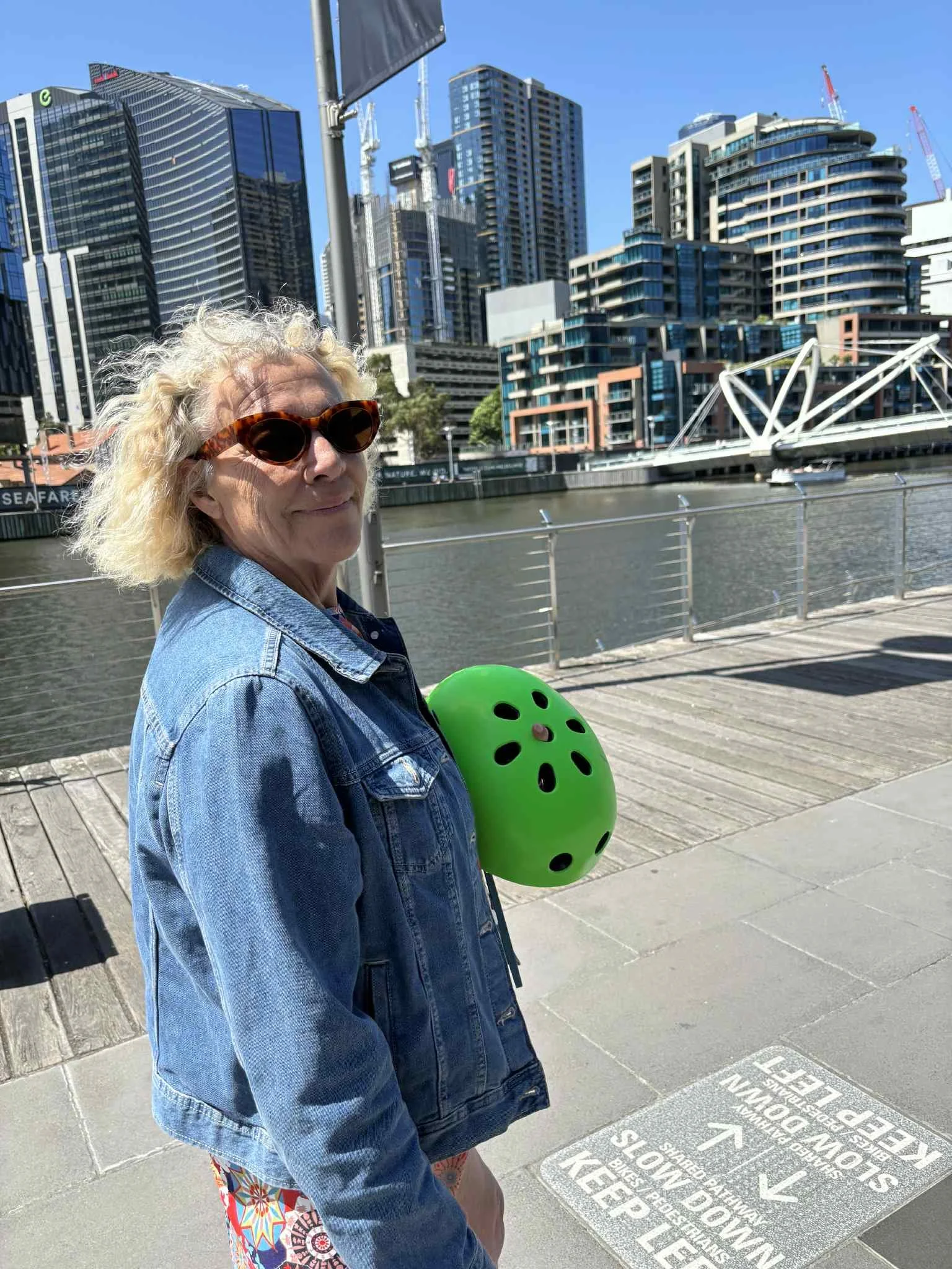 Woman with blonde curly hair wearing sunglasses and denim jacket standing on a riverside walkway in an urban setting with tall buildings and construction cranes in the background.