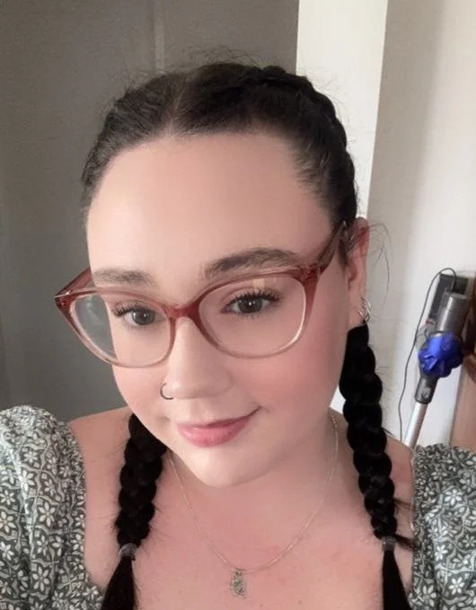A young woman with braided dark hair, wearing glasses, a nose ring, and a patterned top, smiling at the camera indoors.
