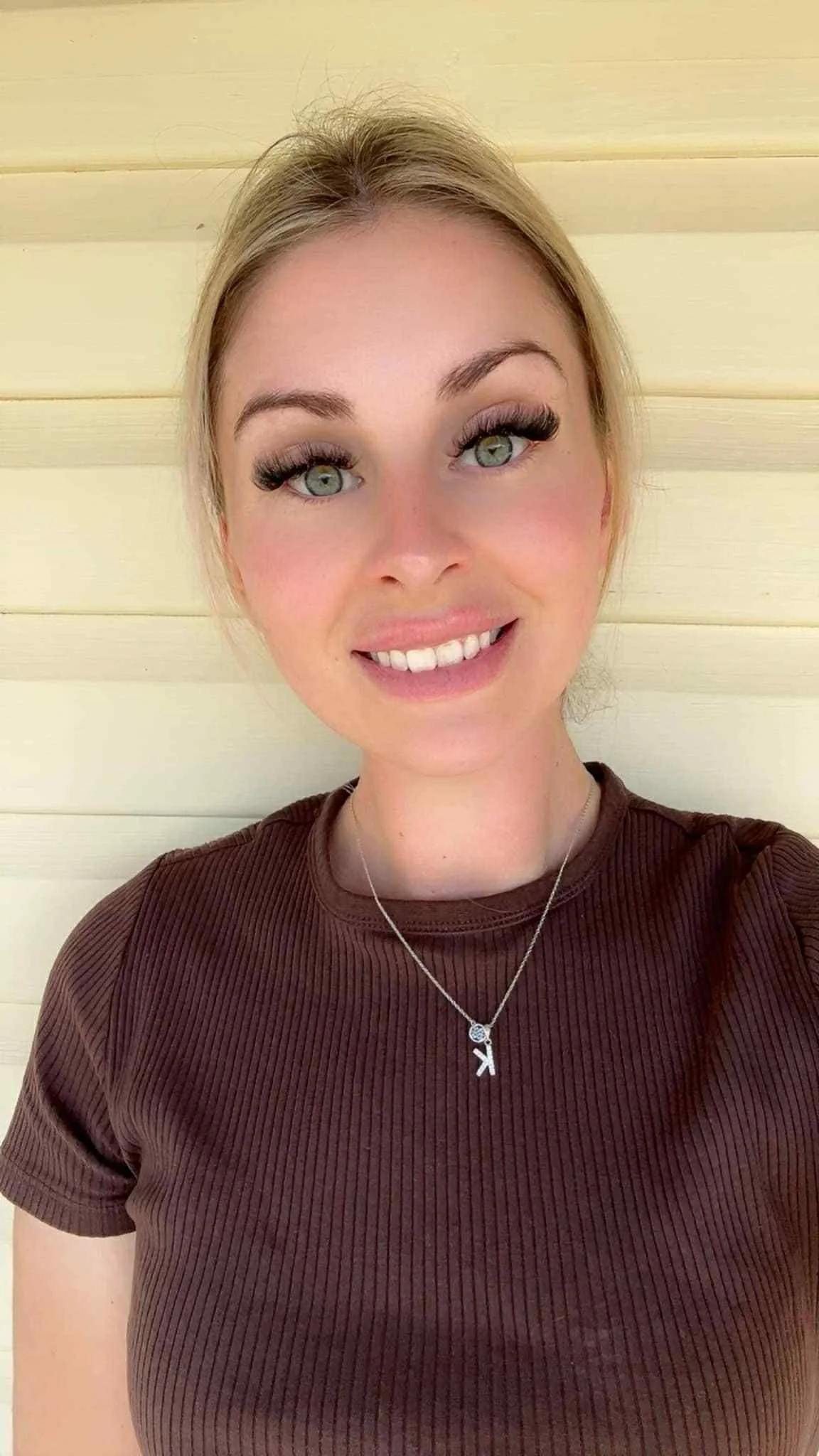A woman with blonde hair, wearing a brown ribbed shirt and a necklace with a letter K pendant, smiling in front of a cream-colored wooden wall.