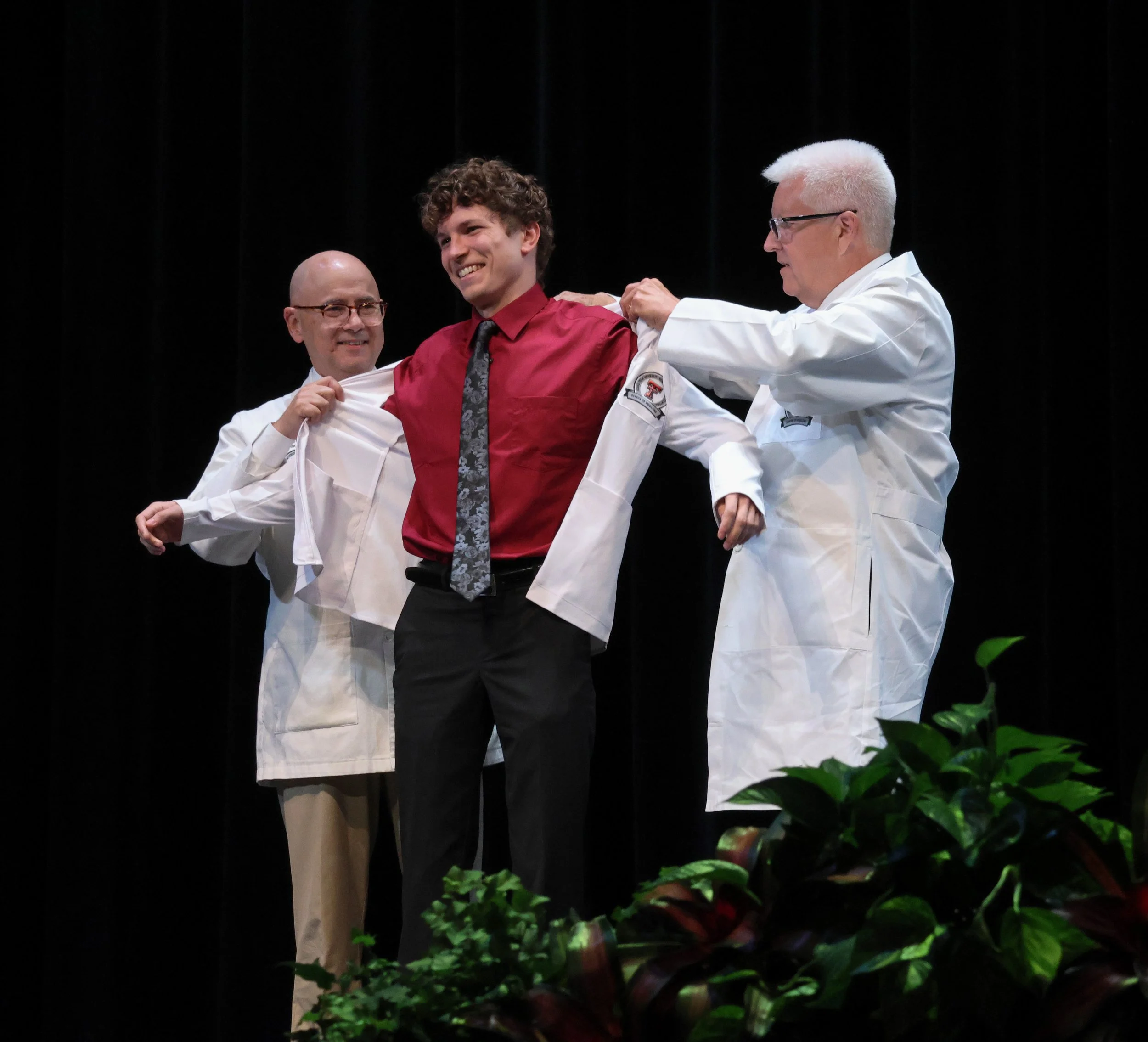 Dr. John DeToledo, dean of the School of Medicine at Texas Tech University Health Sciences Center, and Dr. Brian Schroeder help vest medical student Derek Huber of Keller, Texas, into his white coat during the White Coat Ceremony on July 25, 2025 in