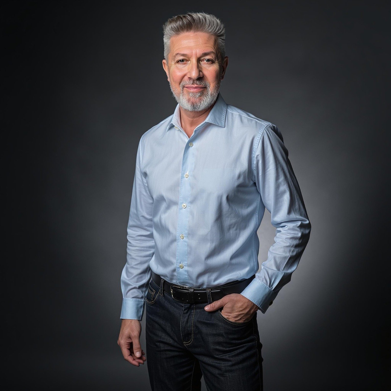 Hombre de cabello gris y barba sonriendo, vestido con camisa de vestir azul y jeans, de fondo oscuro.