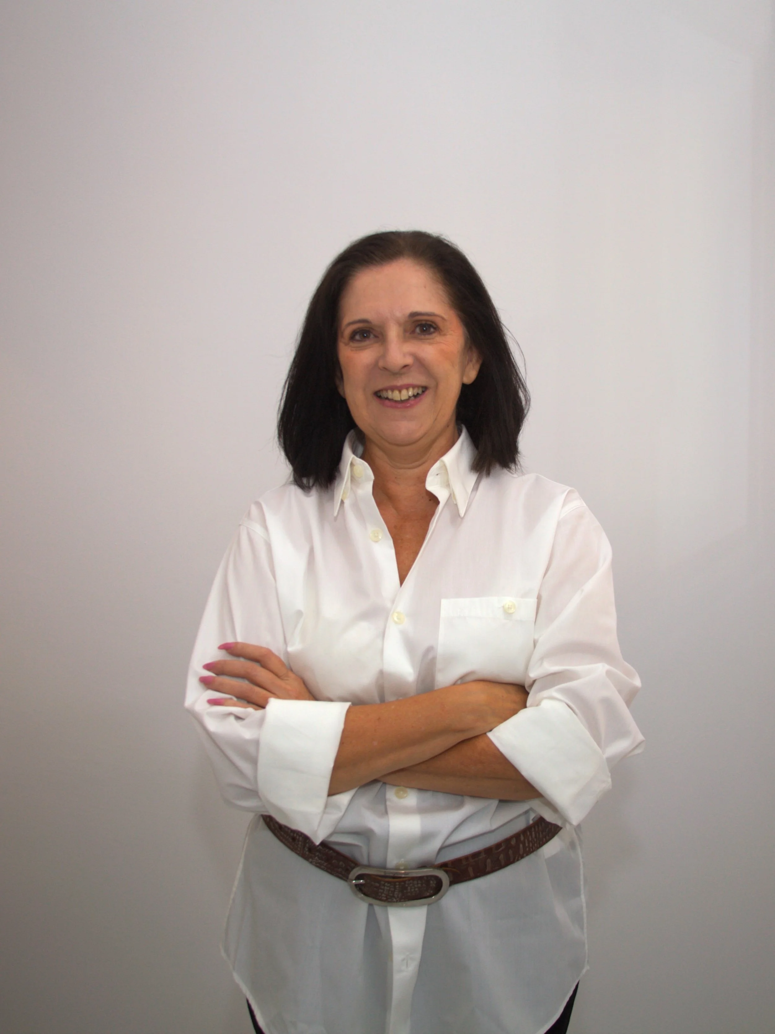 Mujer de cabello oscuro, camiseta blanca y pantalón blanco con cinturón marrón, con brazos cruzados, sonriendo frente a una pared blanca.