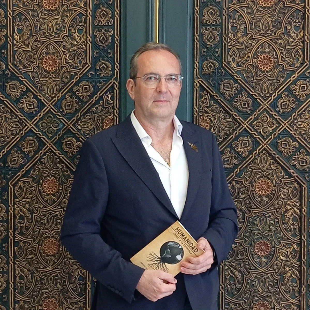 Hombre con gafas, ropa formal, sosteniendo un libro titulado 'Humanidad'. Fondo decorativo con patrón intrincado en tonos dorados y azules.