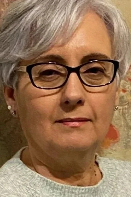 Retrato de una mujer de cabello canoso, usa gafas negras y aretes plateados, con fondo de pared decorada con flores.