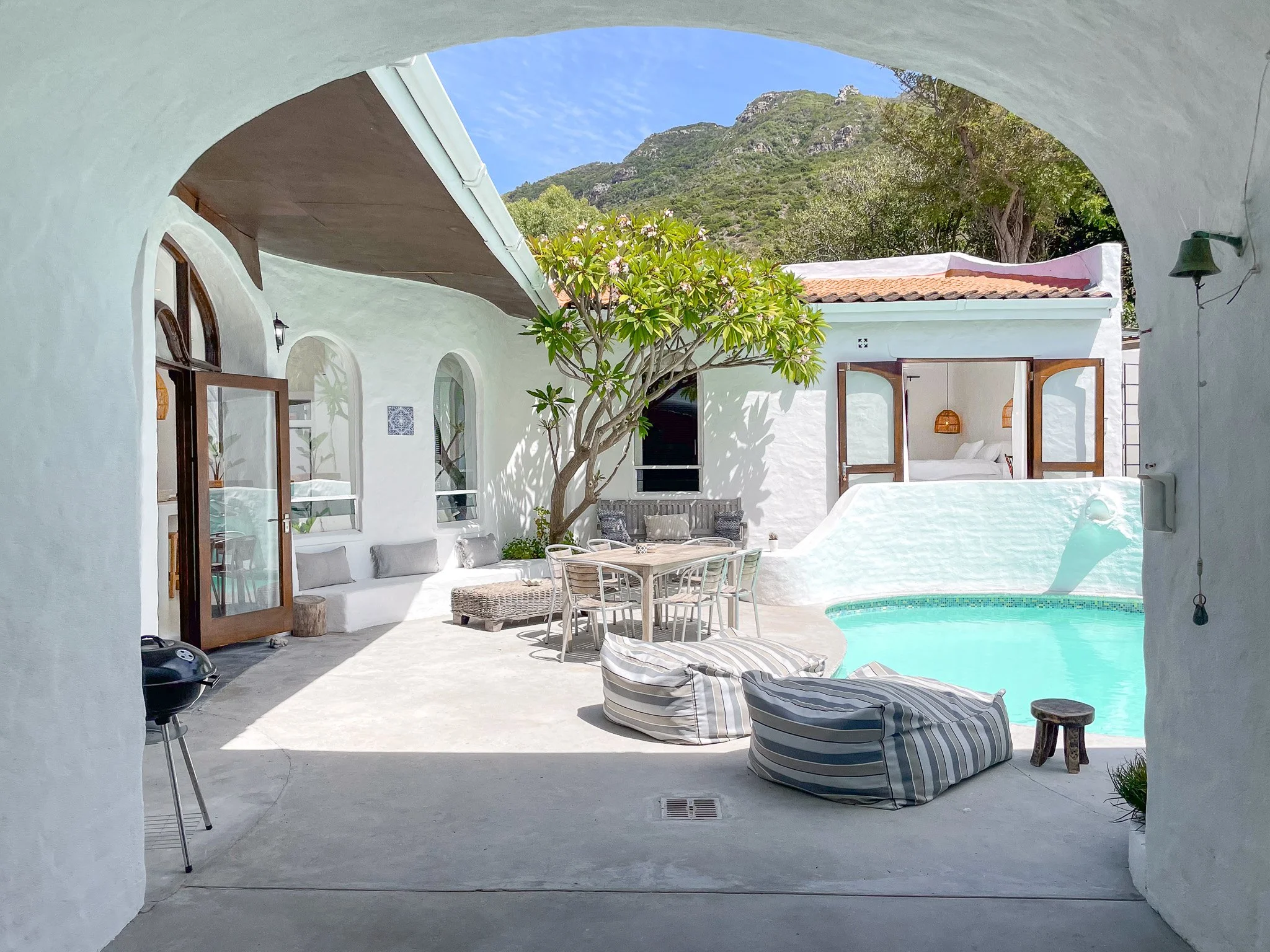 Bright living room with white brick walls, large windows with white curtains, wooden coffee table, and various armchairs with cushions, open to a patio area.