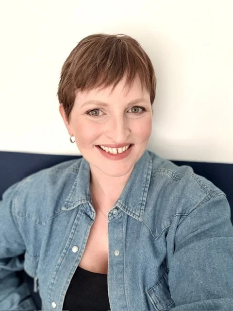 A woman with short reddish-brown hair smiling, wearing a denim jacket and earrings, sitting against a light background.