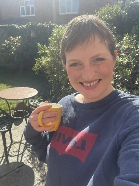 A woman with short hair smiling and holding a yellow mug in a backyard garden on a sunny day.
