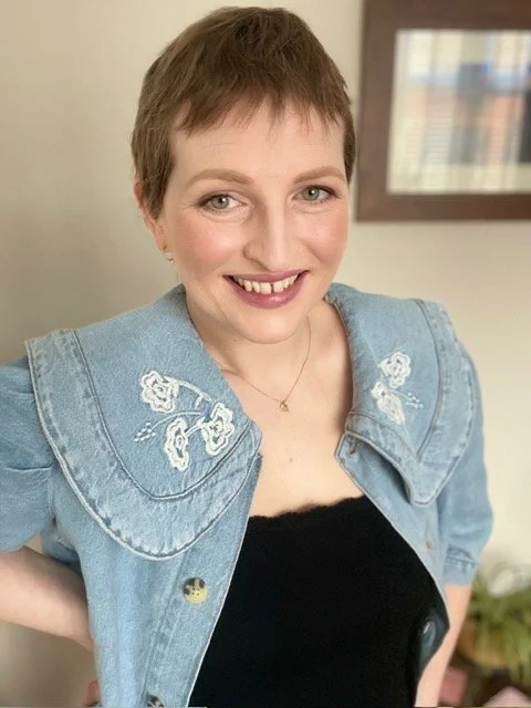 A woman with short brown hair smiling, wearing a black top and a blue denim jacket with embroidered white flowers on the shoulders, indoors.