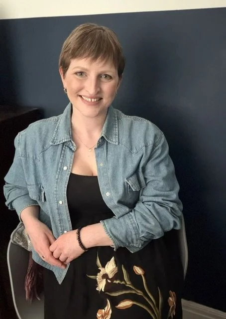 A woman smiling, sitting on a chair, wearing a black dress with floral embroidery and a denim jacket, with short brown hair, in an indoor setting.