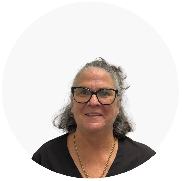 A middle-aged woman with curly gray hair wearing black glasses, gold earrings, and a gold chain necklace, smiling at the camera against a plain white background.