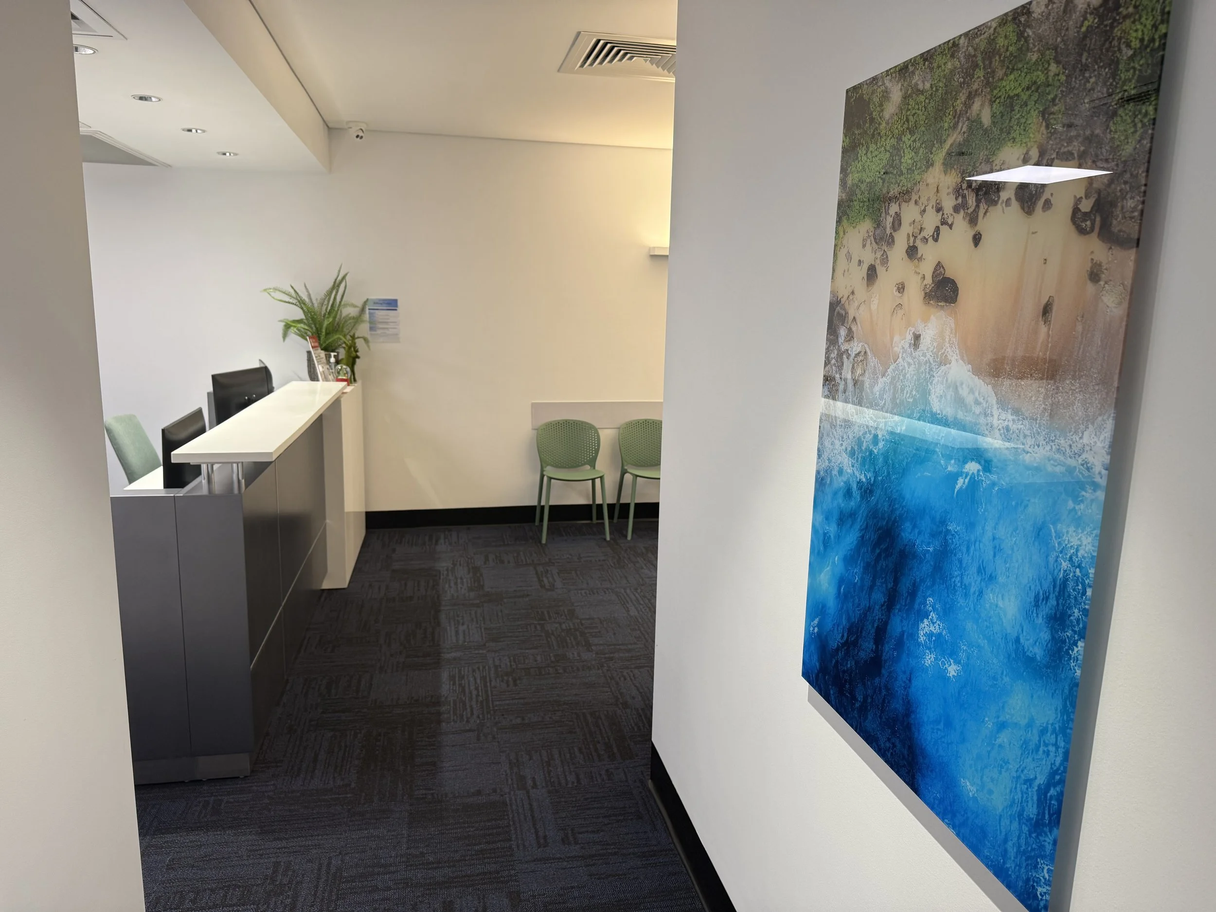 Interior of an office with security cameras on the ceiling, a white reception desk with black and green chairs, a plant, and a large ocean-themed painting on the wall.