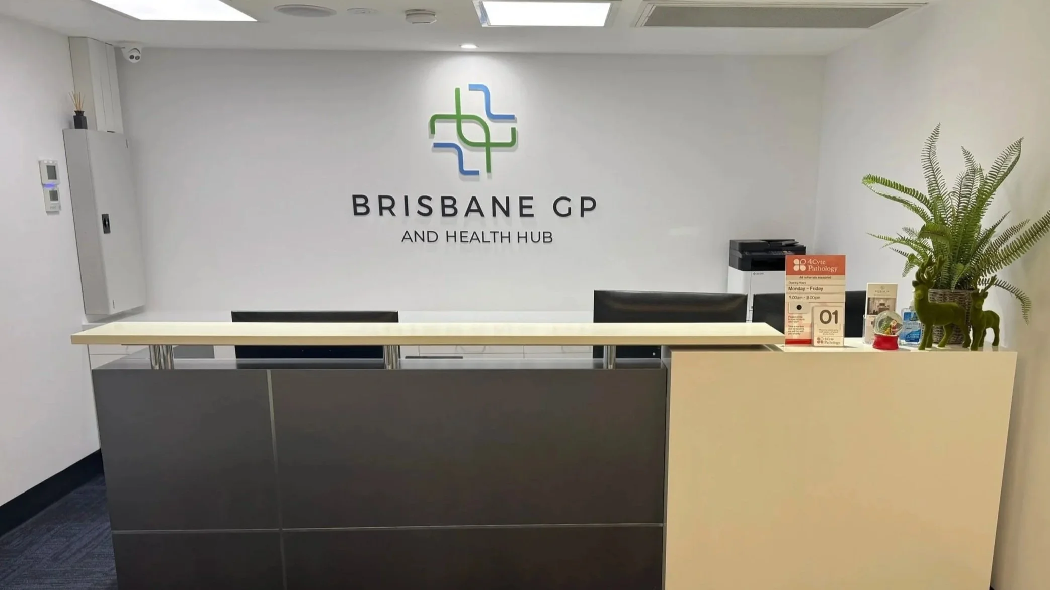 Reception area at Brisbane GP and Health Hub with a desk, potted plant, and health-related signage.