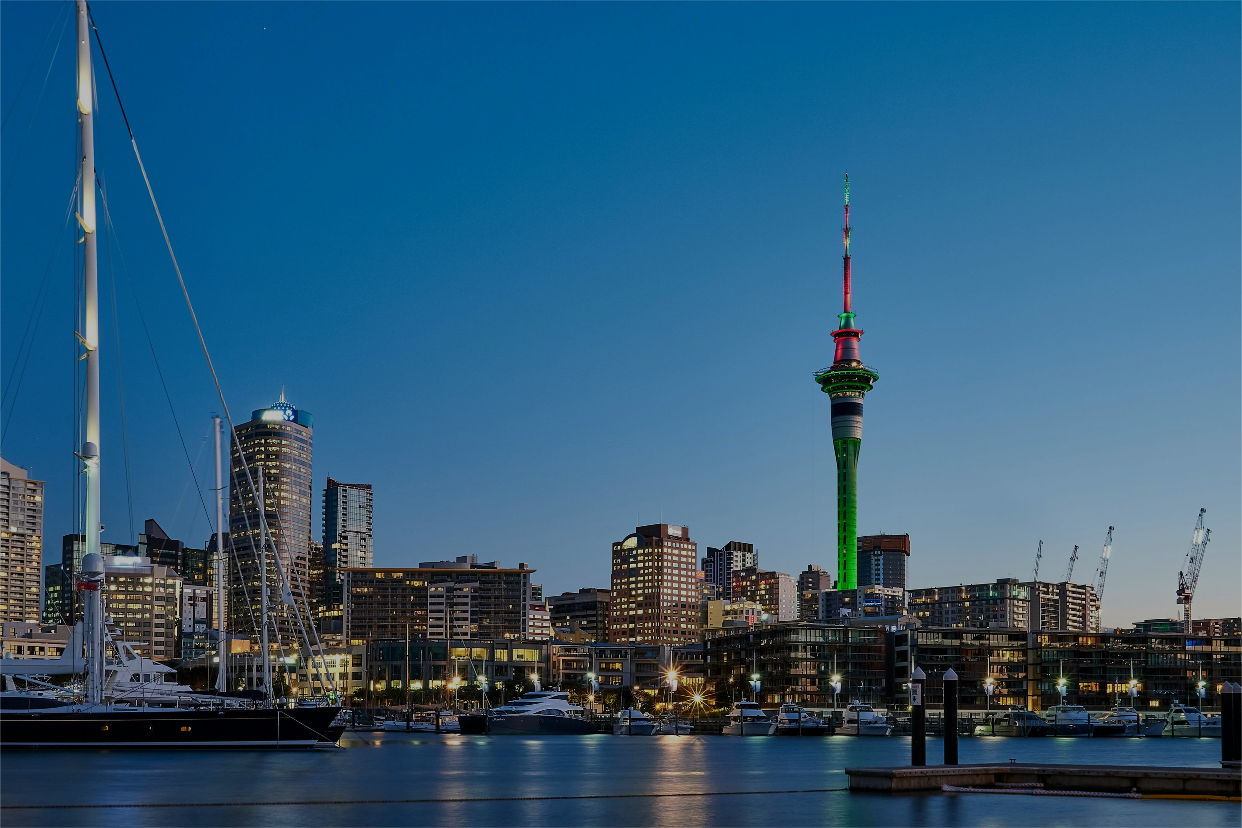 Auckland, Job agency, try out careers, Nighttime cityscape featuring the Sky Tower with green and red lights, high-rise buildings, and a marina with boats and yachts in the foreground.