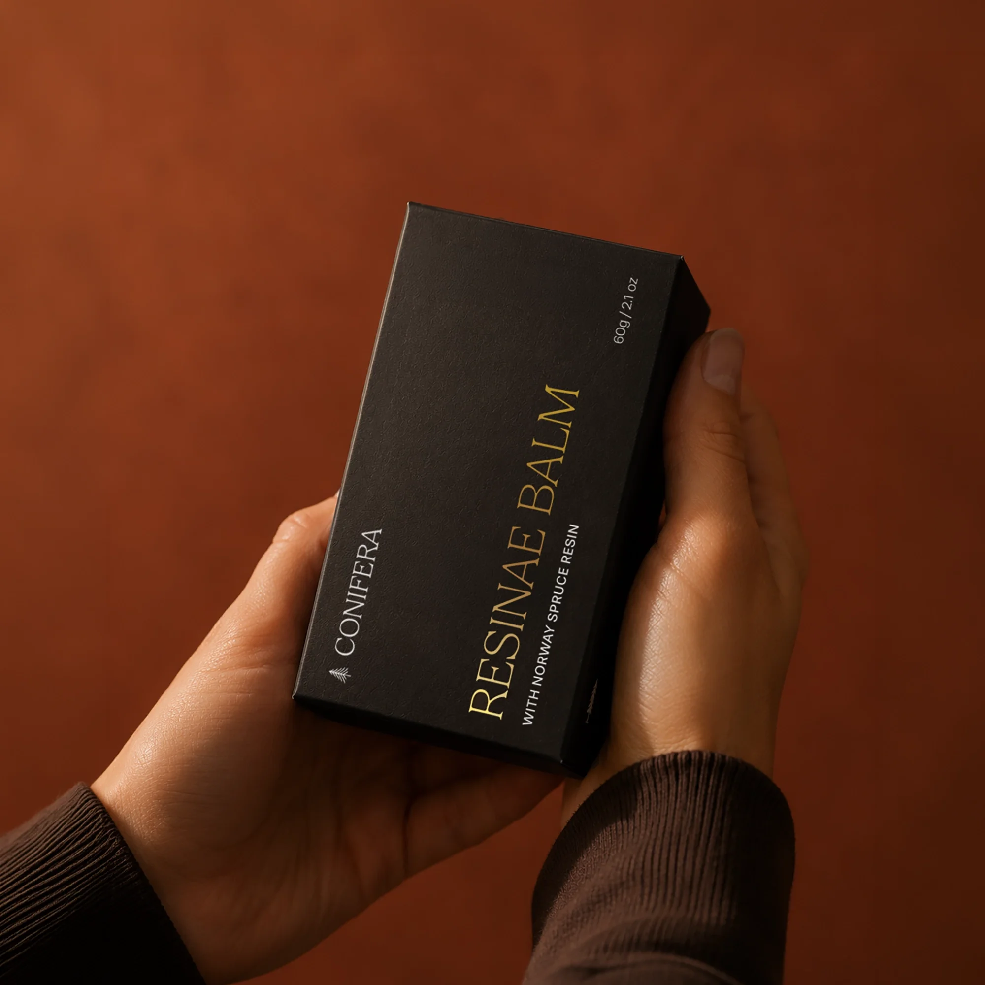 Hands holding a black Conifera Resinae Balm box with gold serif typography, featuring Norway spruce resin.