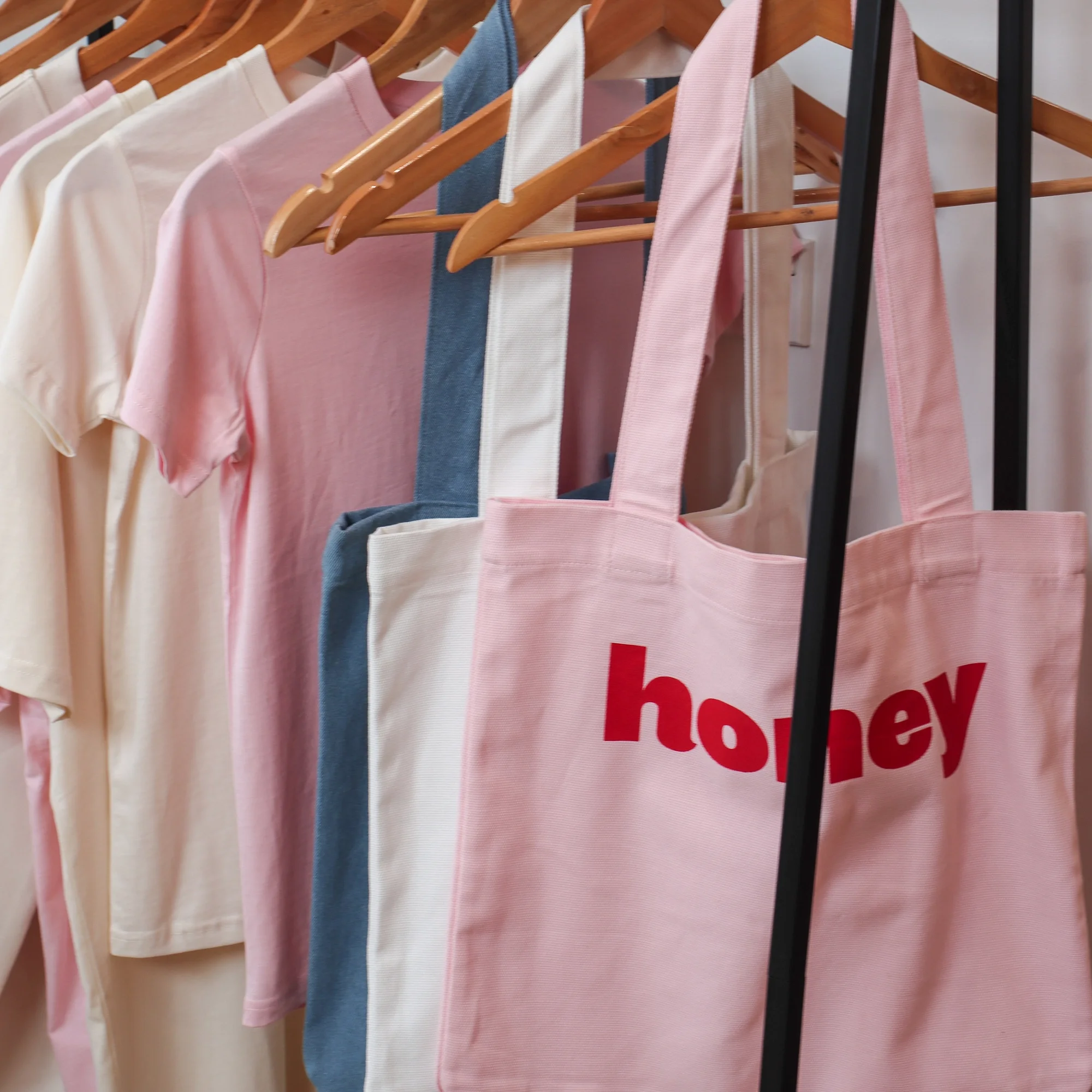 Close-up of Honey branded t-shirts in cream, pink, and blue on wooden hangers, with a pink tote bag featuring the red Honey logo.