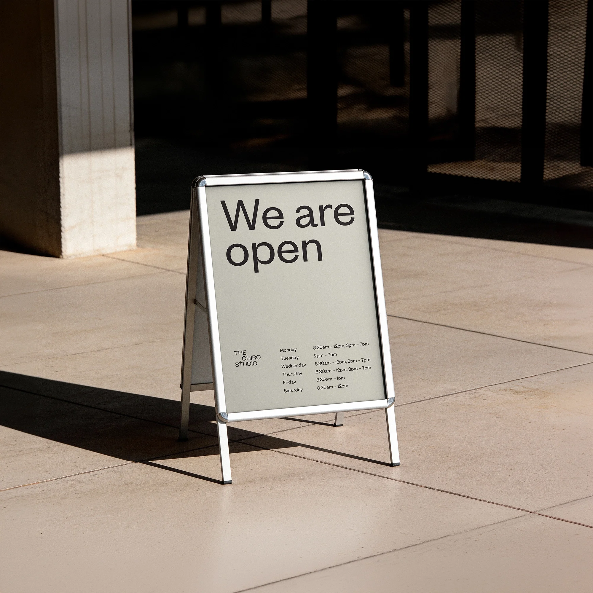 The Chiro Studio sandwich board sign reading 'We are open' with weekly opening hours listed, standing on a sunlit concrete footpath.