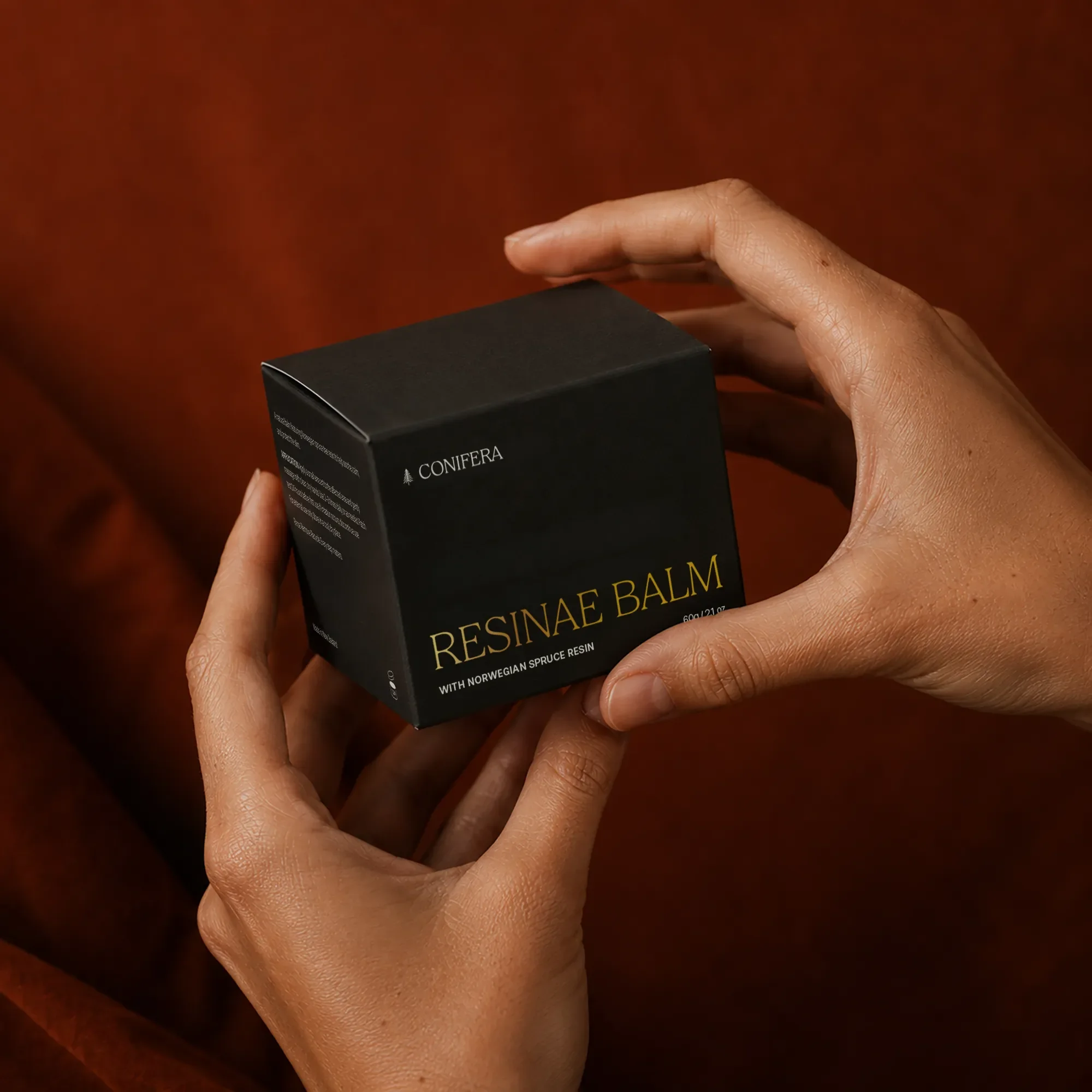 Hands holding a black Conifera Resinae Balm box with gold serif typography, set against a rust-orange velvet backdrop.