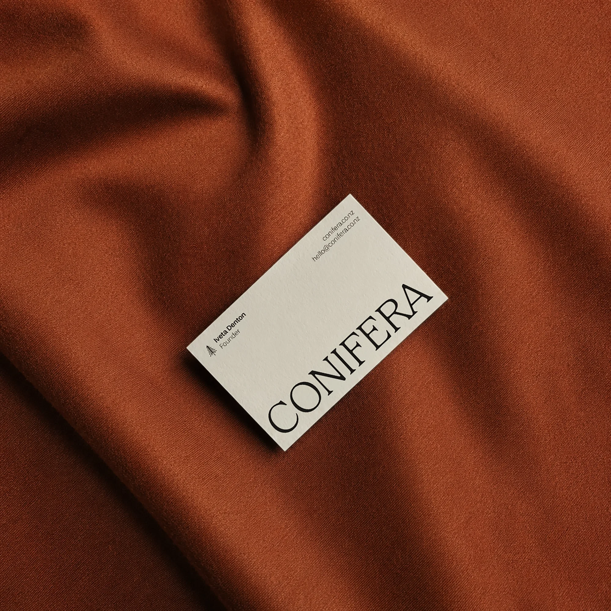 Conifera business card with serif logo and pine tree icon, styled on rust-orange silk fabric.
