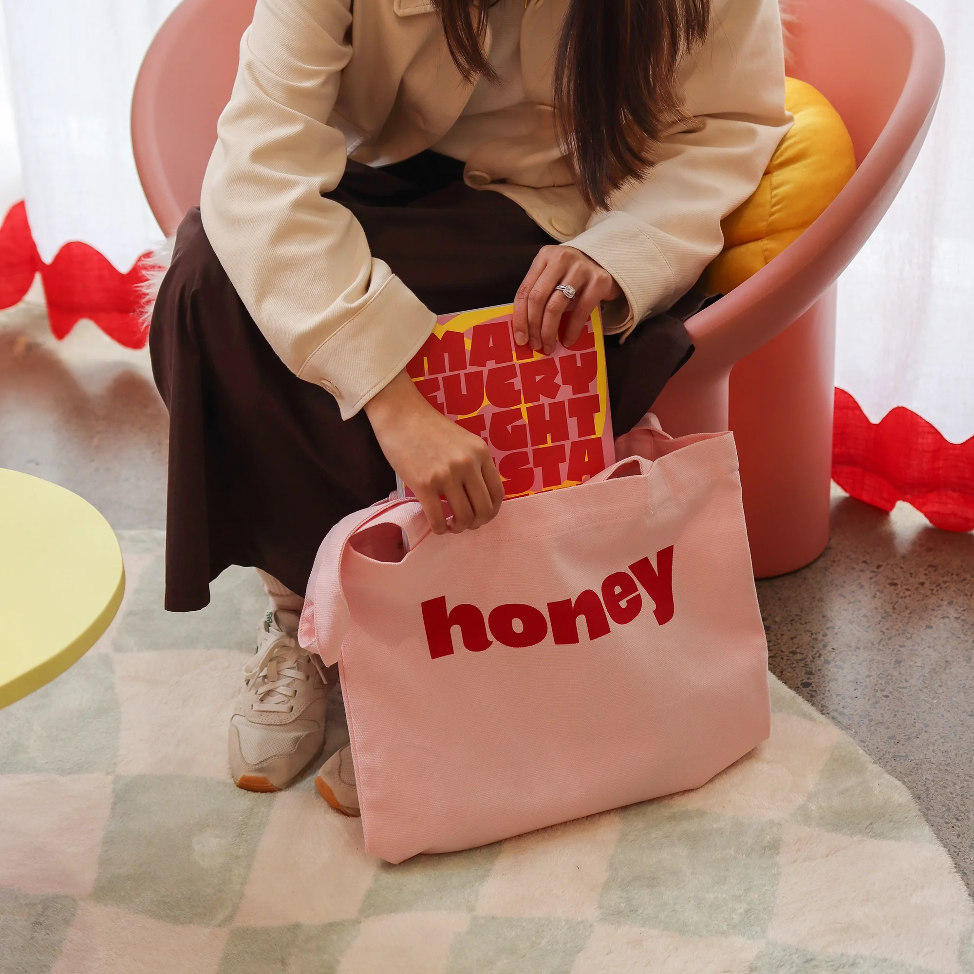 Woman holding a Honey branded pink tote bag with a magazine tucked inside, seated in a pink chair with red wavy trim in the background.