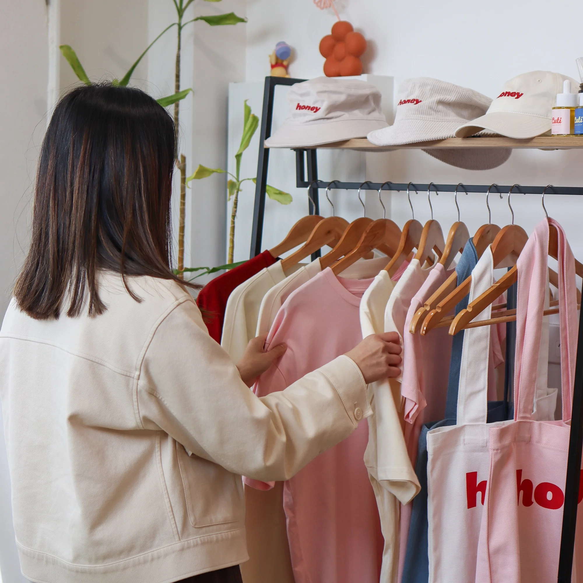 Woman browsing a rack of Honey branded pink, cream, and blue t-shirts and tote bags, with embroidered bucket hats and caps displayed on the shelf above.