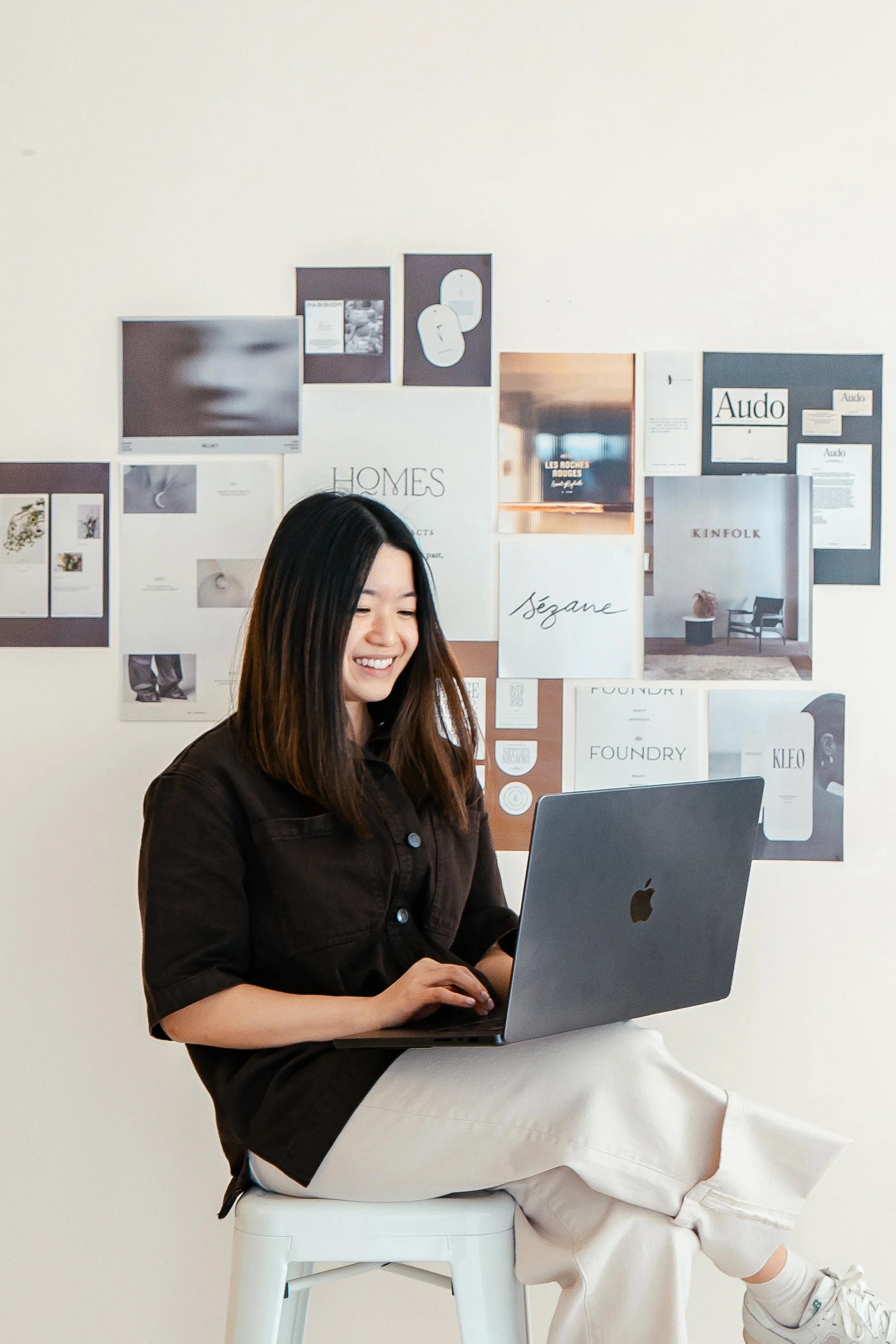 Wonder Studio founder Joyce O'Riley smiling while working at her laptop, surrounded by a design inspiration board of fashion and lifestyle brands.