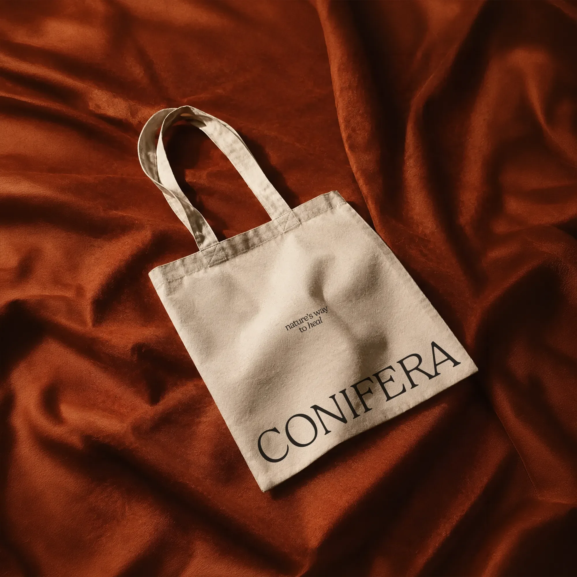 Cream canvas Conifera tote bag with 'nature's way to heal' tagline, laid flat on rust-orange silk fabric.