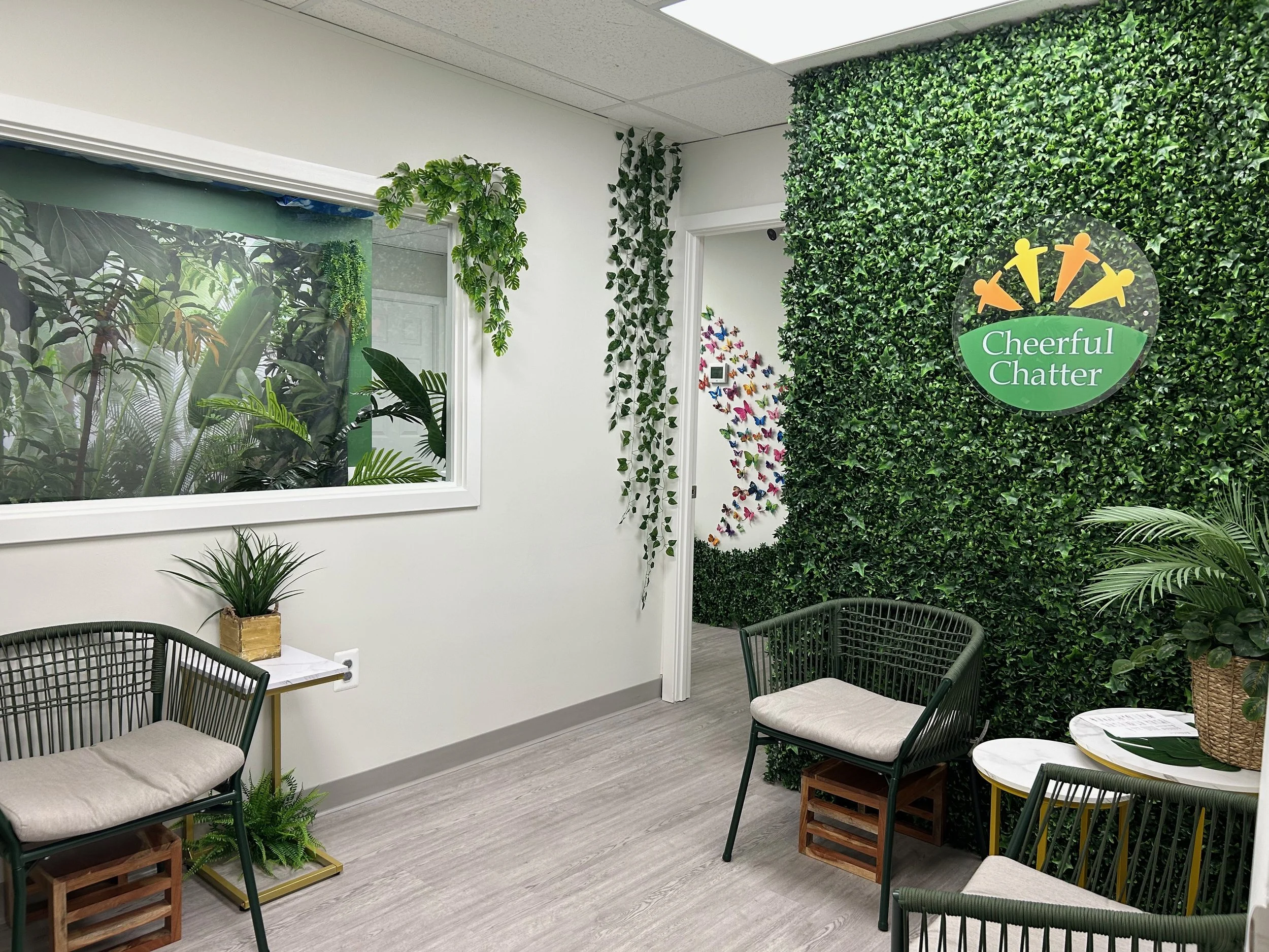 Indoor waiting area with green leafy decor, a sign reading 'Cheerful Chatter', and modern furniture including chairs, tables, and plants.