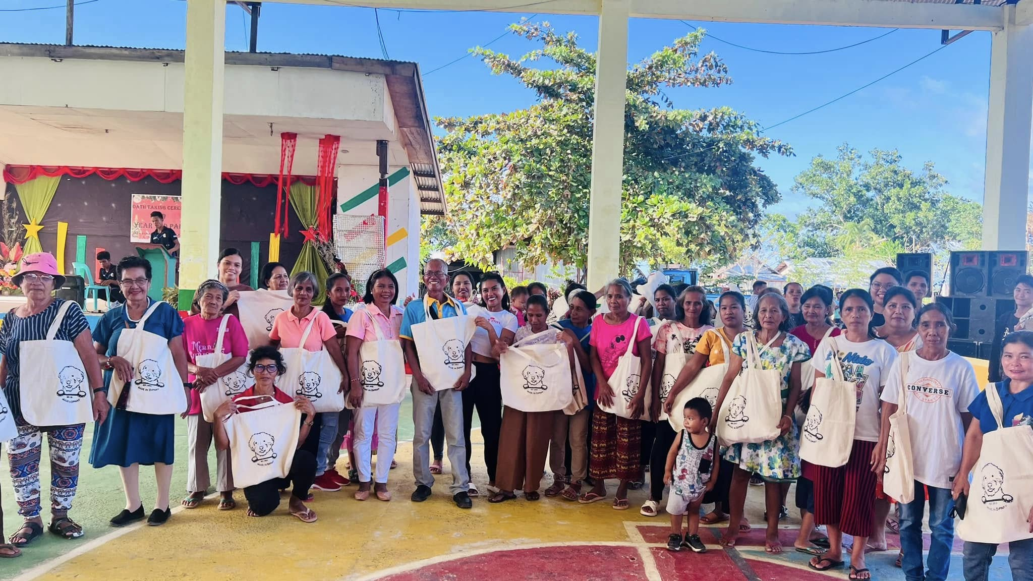 Group of people standing on a colorful outdoor basketball court, holding tote bags with a cartoon dog drawing, under a covered area with a small stage. Trees and a blue sky are in the background.