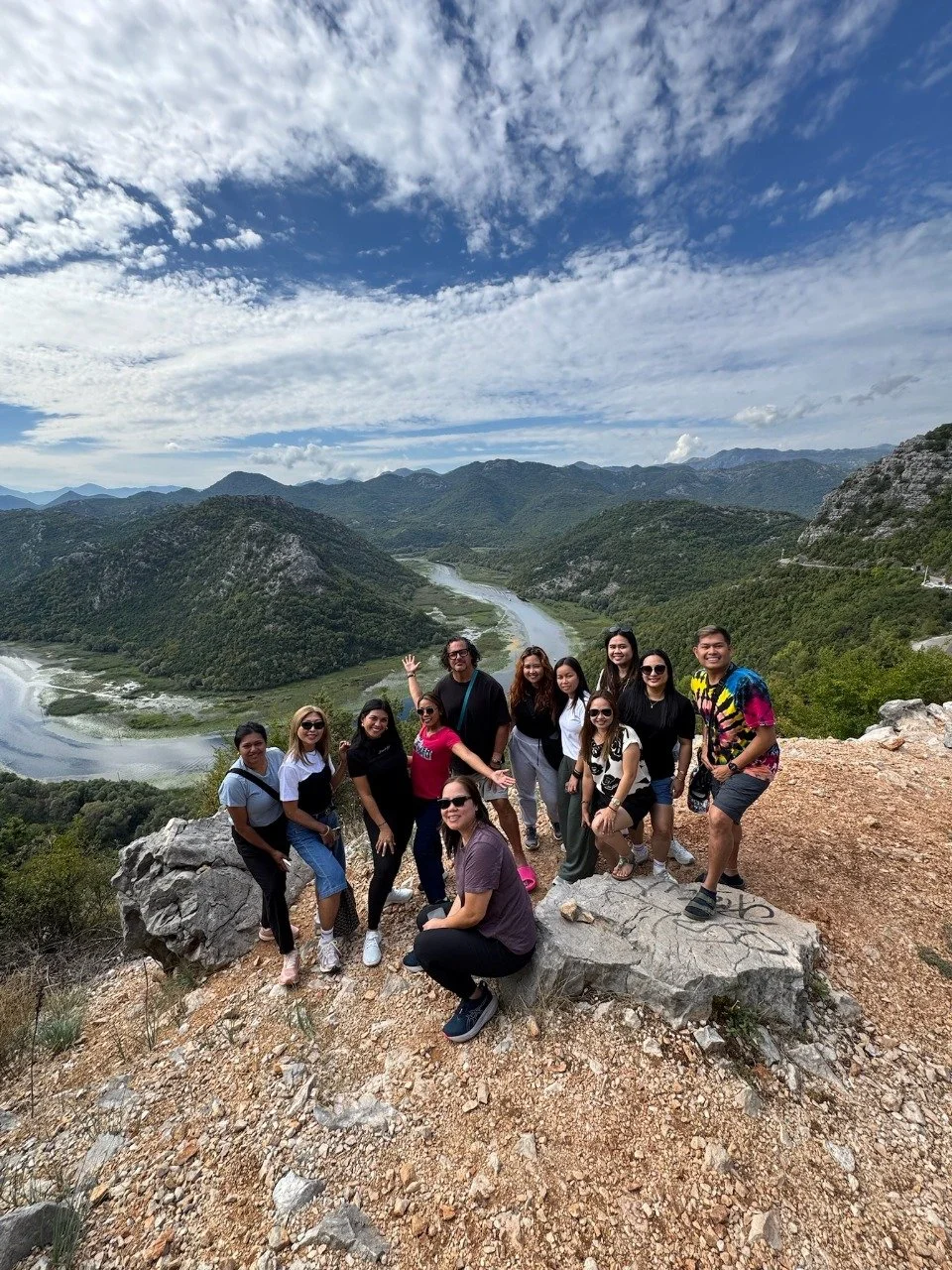 Group of eleven people standing on a rocky overlook with a river and mountain range in the background, under partly cloudy skies in Lake Skadar, Montenegro.