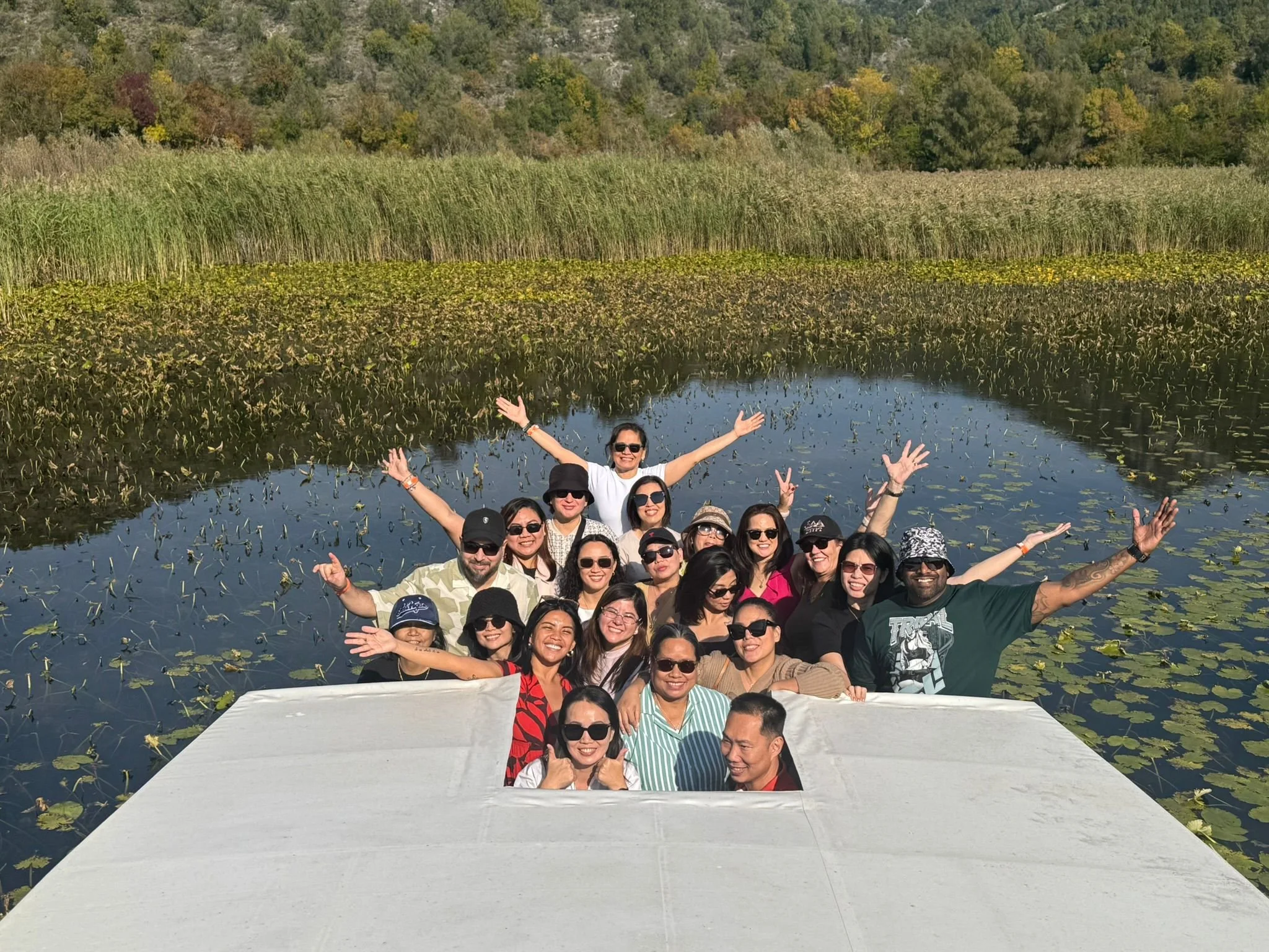 Group of people on a boat in a pond with lily pads, surrounded by tall reeds and trees in the background, enjoying a sunny day.